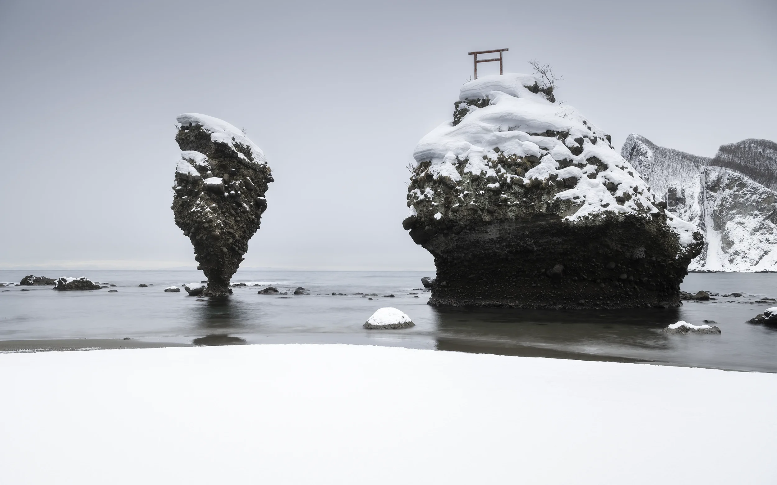 Cliché d'une plage enneigée avec des formations rocheuses, dont deux grandes et une petite en premier plan, et un torii rouge au sommet de la falaise à droite.