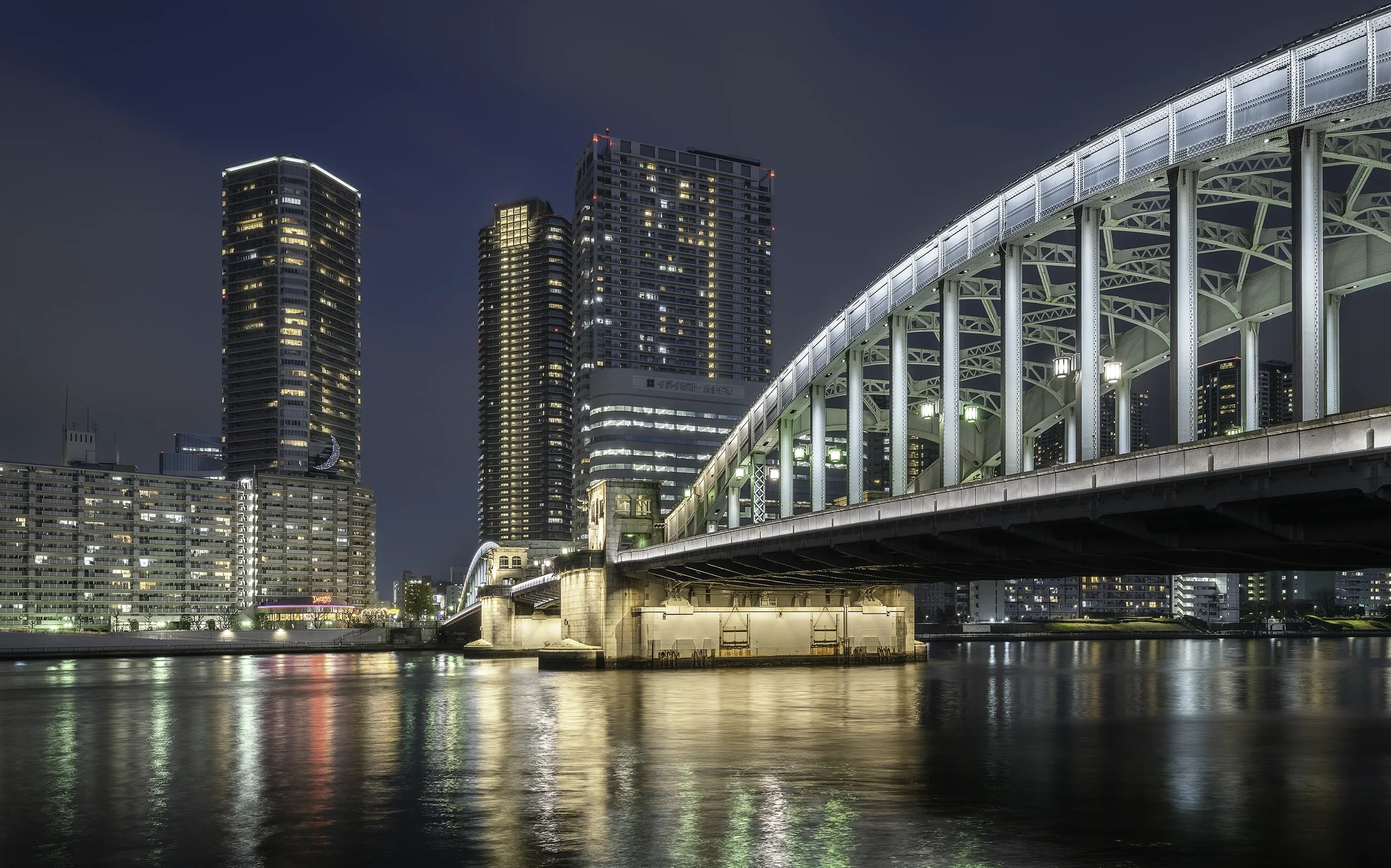 Ville moderne avec des gratte-ciel illuminés, un pont métallique éclairé au bord d'une rivière la nuit, reflection dans l'eau