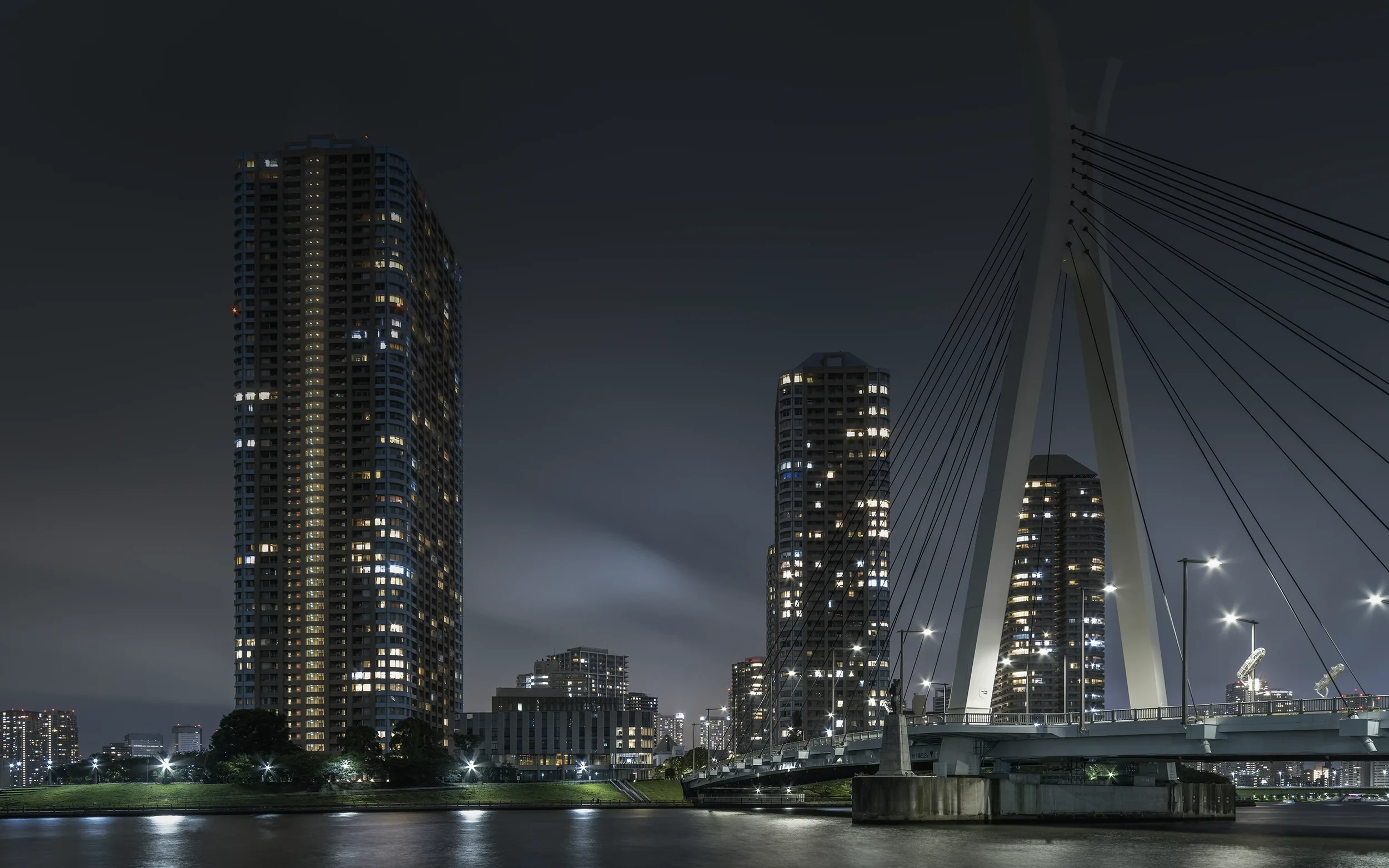 Vue nocturne d'une ville avec des gratte-ciel éclairés et un pont piétonnier moderne au premier plan, reflété dans l'eau.