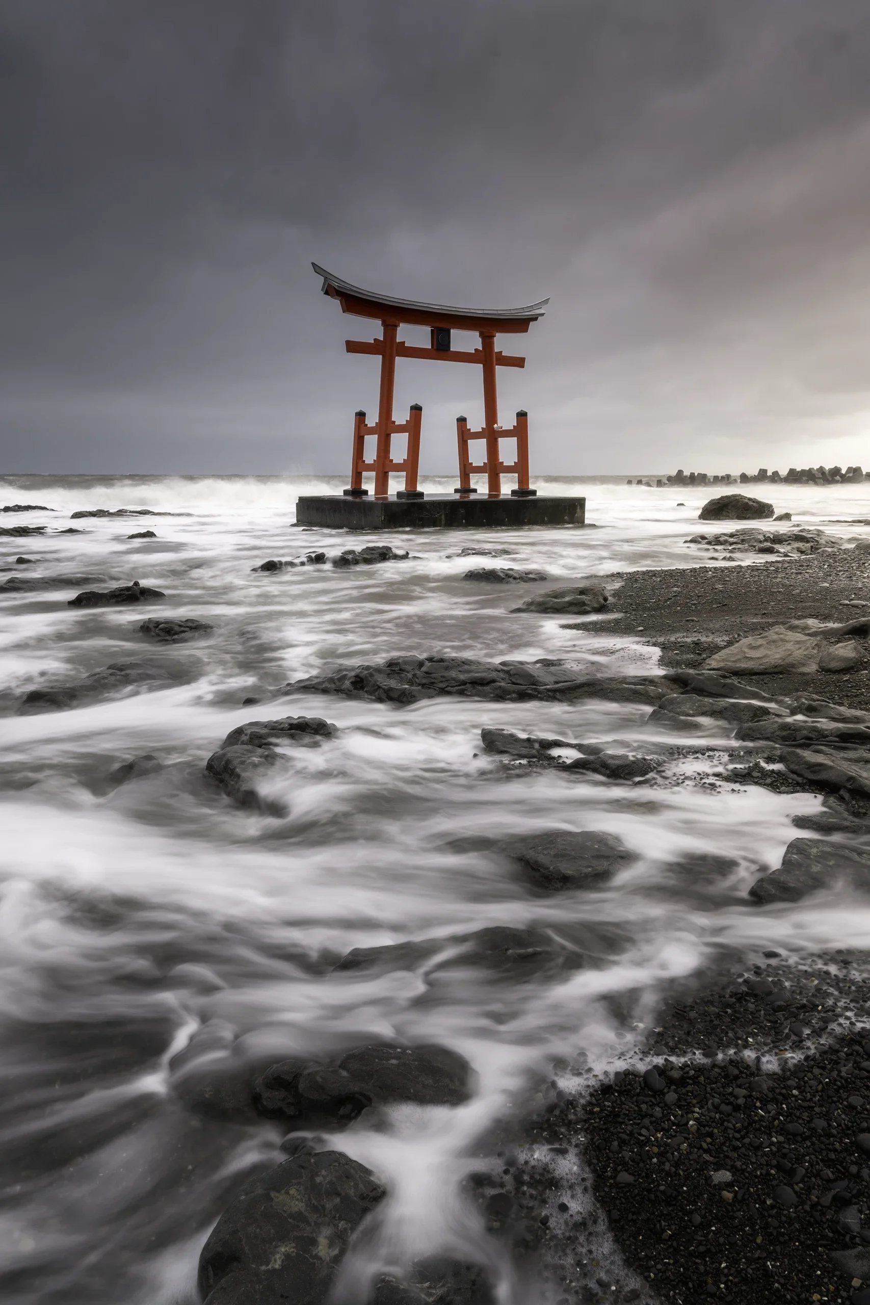 Une statue torii rouge située dans la mer, avec un ciel nuageux en arrière-plan et des rochers autour de la côte.