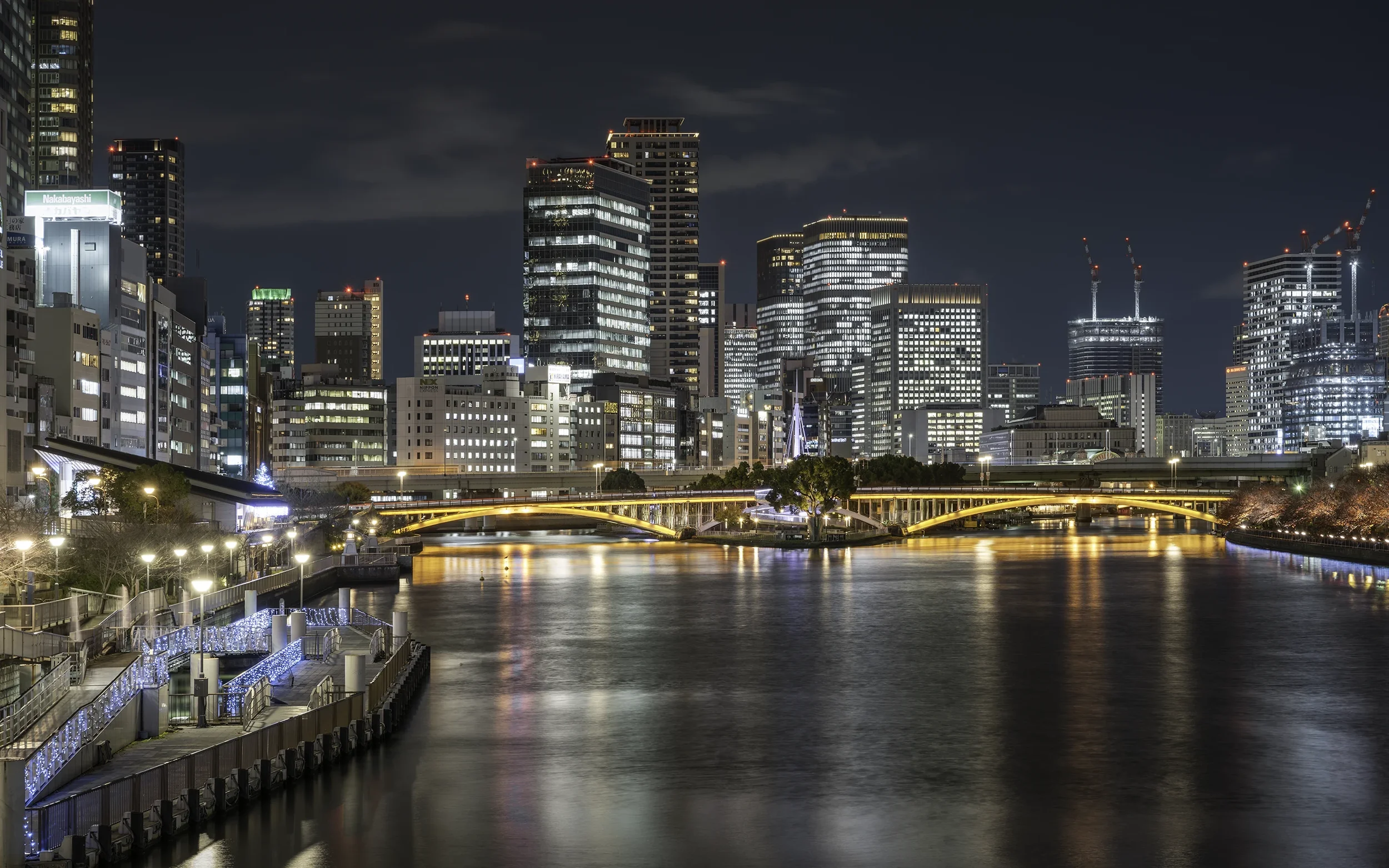 Vue nocturne d'une ville moderne avec beaucoup de gratte-ciel illuminés, un fleuve au premier plan avec reflet des lumières, un pont éclairé en arc, et des bâtiments en construction avec grues.