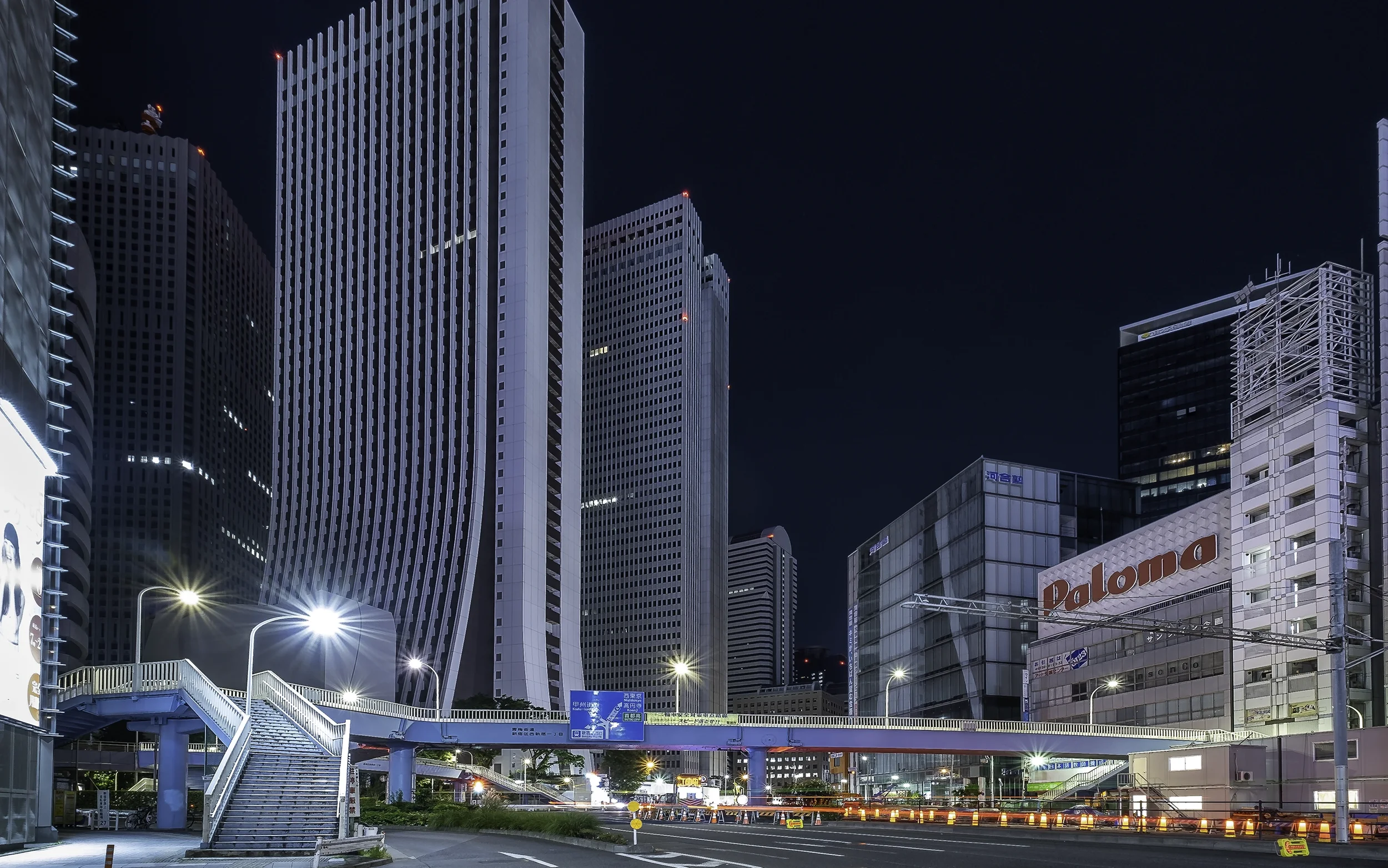 Vue nocturne d'une ville avec plusieurs gratte-ciel modernes, un pont piétonnier bleu en avant-plan, et des lampadaires lumineux.