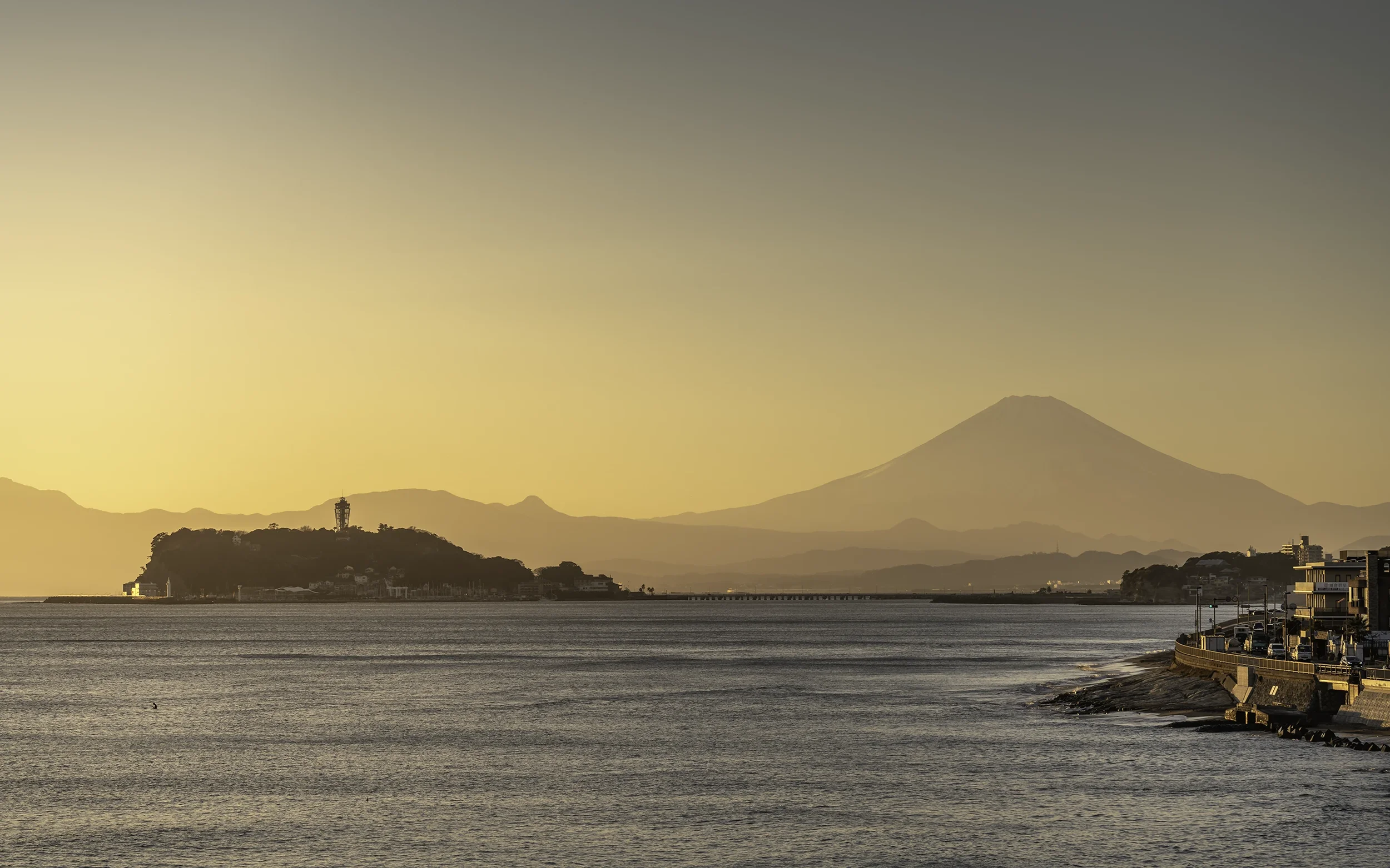 Coucher de soleil sur la mer avec une île au centre et une montagne au fond, avec des bâtiments en bord de mer à droite.