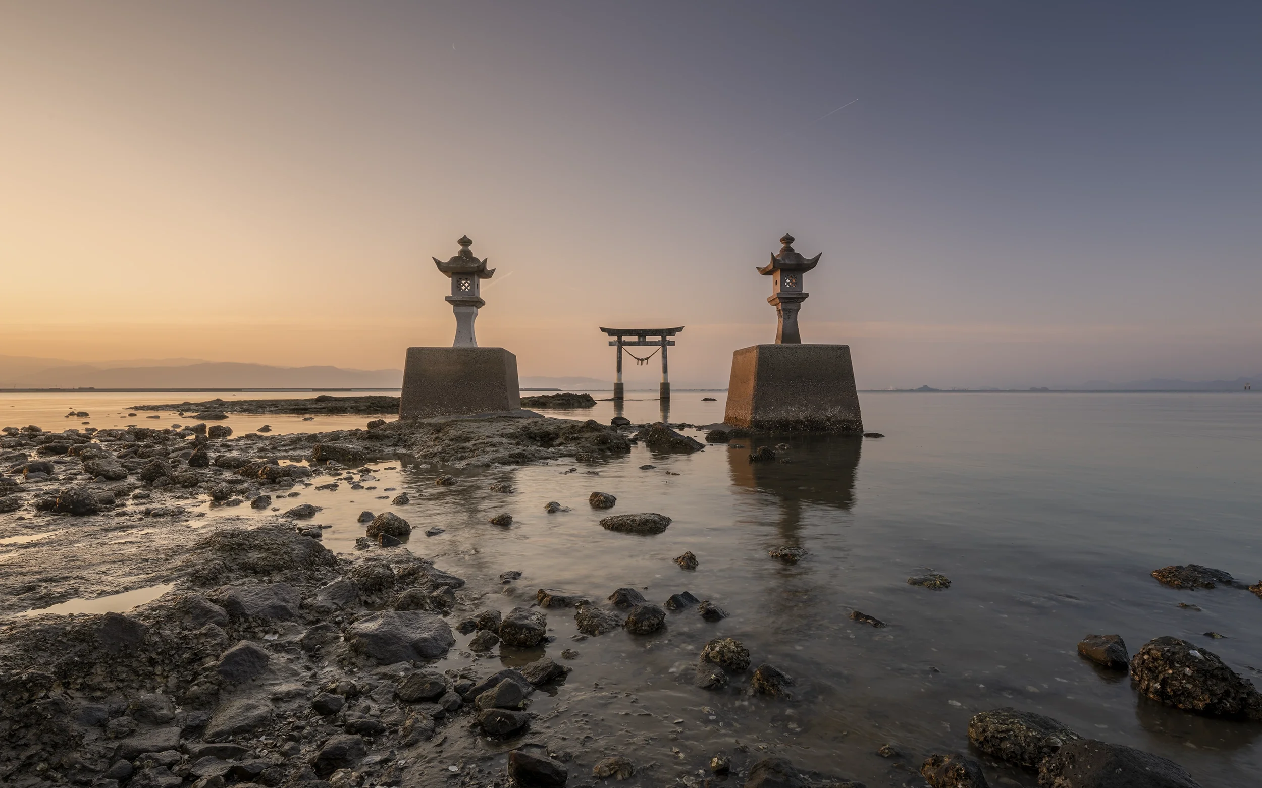 Torii traditionnel japonais dans l'eau au coucher du soleil, avec deux lanternes en pierre sur chaque côté, rocheuse en premier plan.