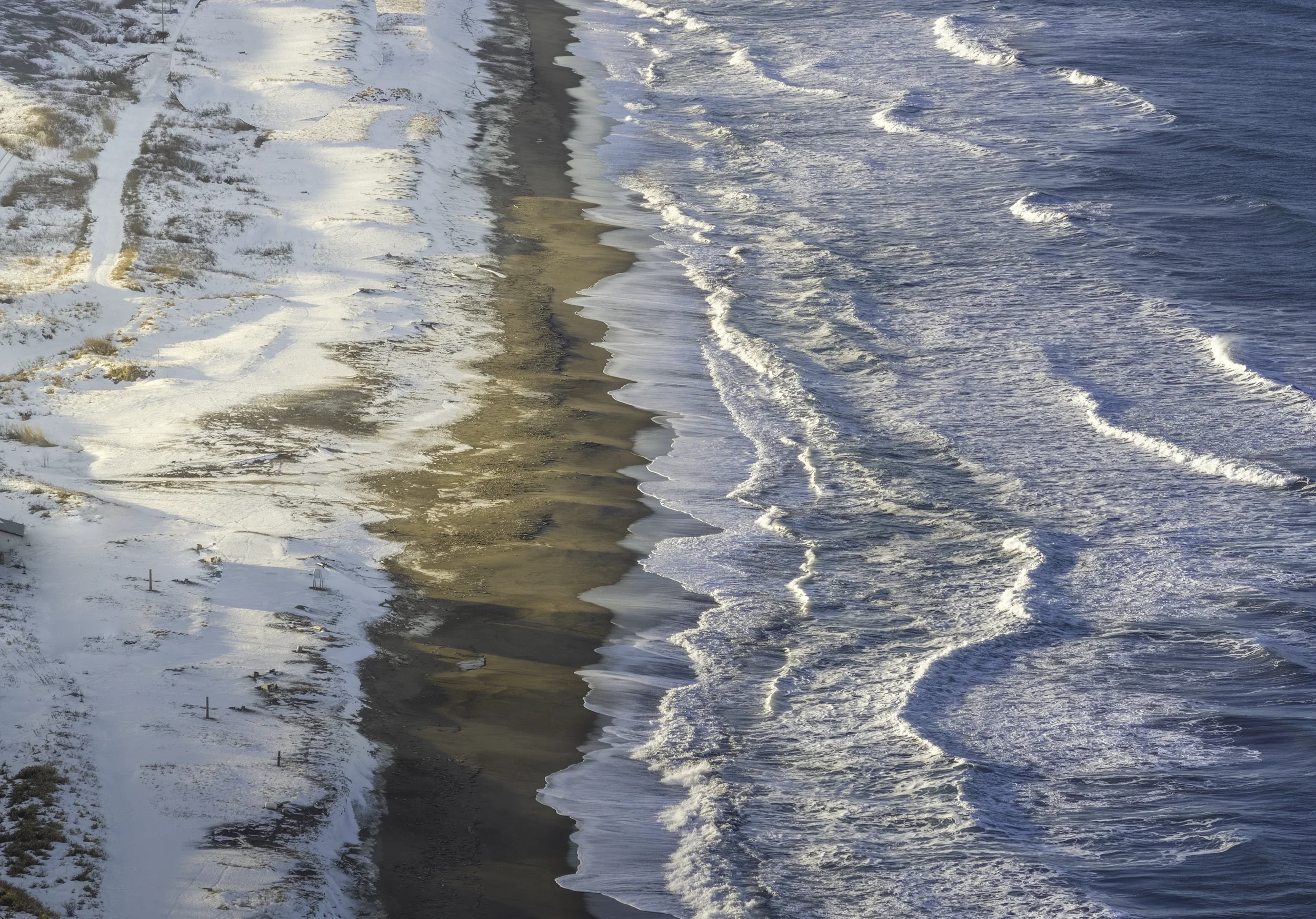 Snow-covered beach along the Sea of Japan in Hokkaido, photographed from above as winter waves create abstract patterns along the coastline.