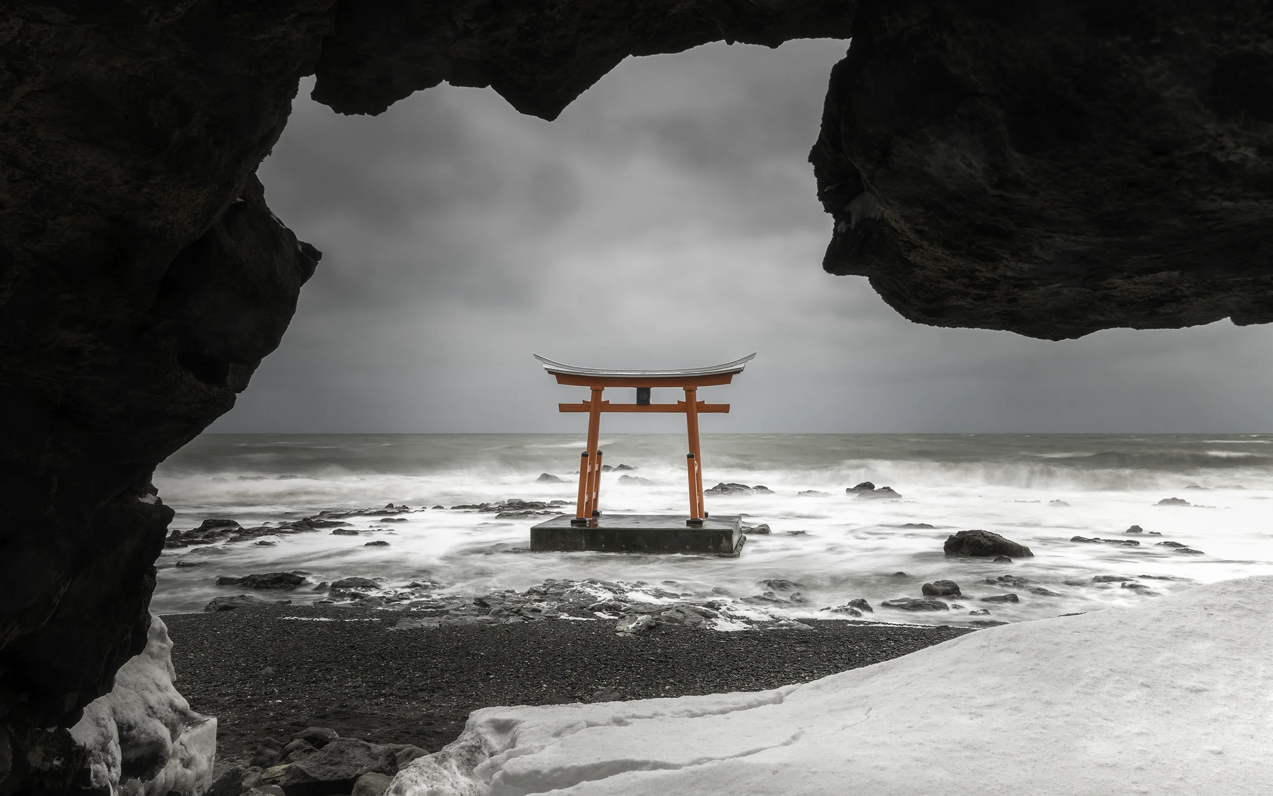 Une sculpture Torii rouge située sur une plateforme dans la mer, vue depuis une grotte sur une plage rocheuse sous un ciel nuageux.