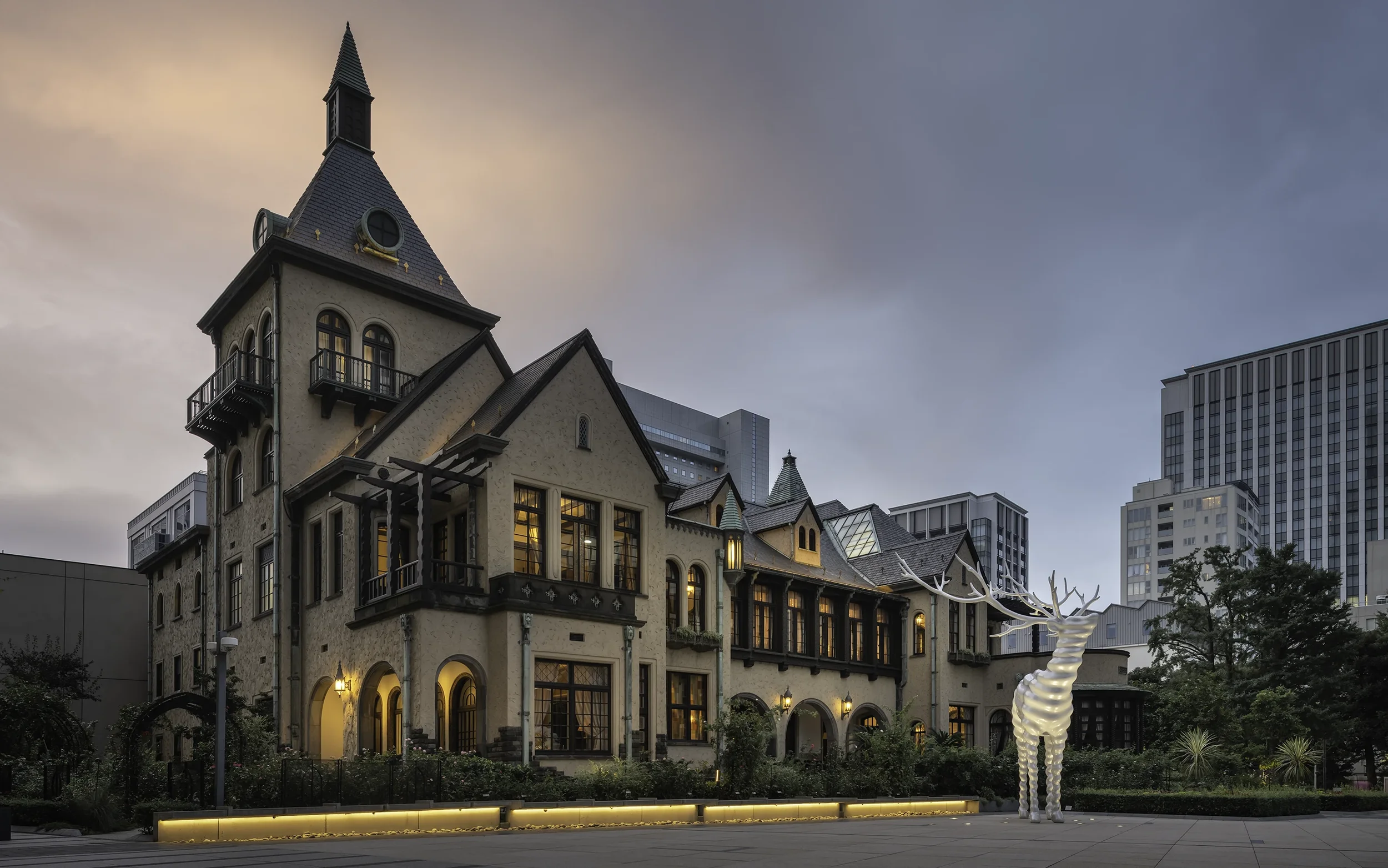 Une grande maison de style castle avec plusieurs fenêtres illuminées, un toit pointu, et un grand cerf en décoration lumineuse devant, dans un environnement urbain avec des immeubles modernes en arrière-plan. Le ciel est en dégradé avec des teintes r