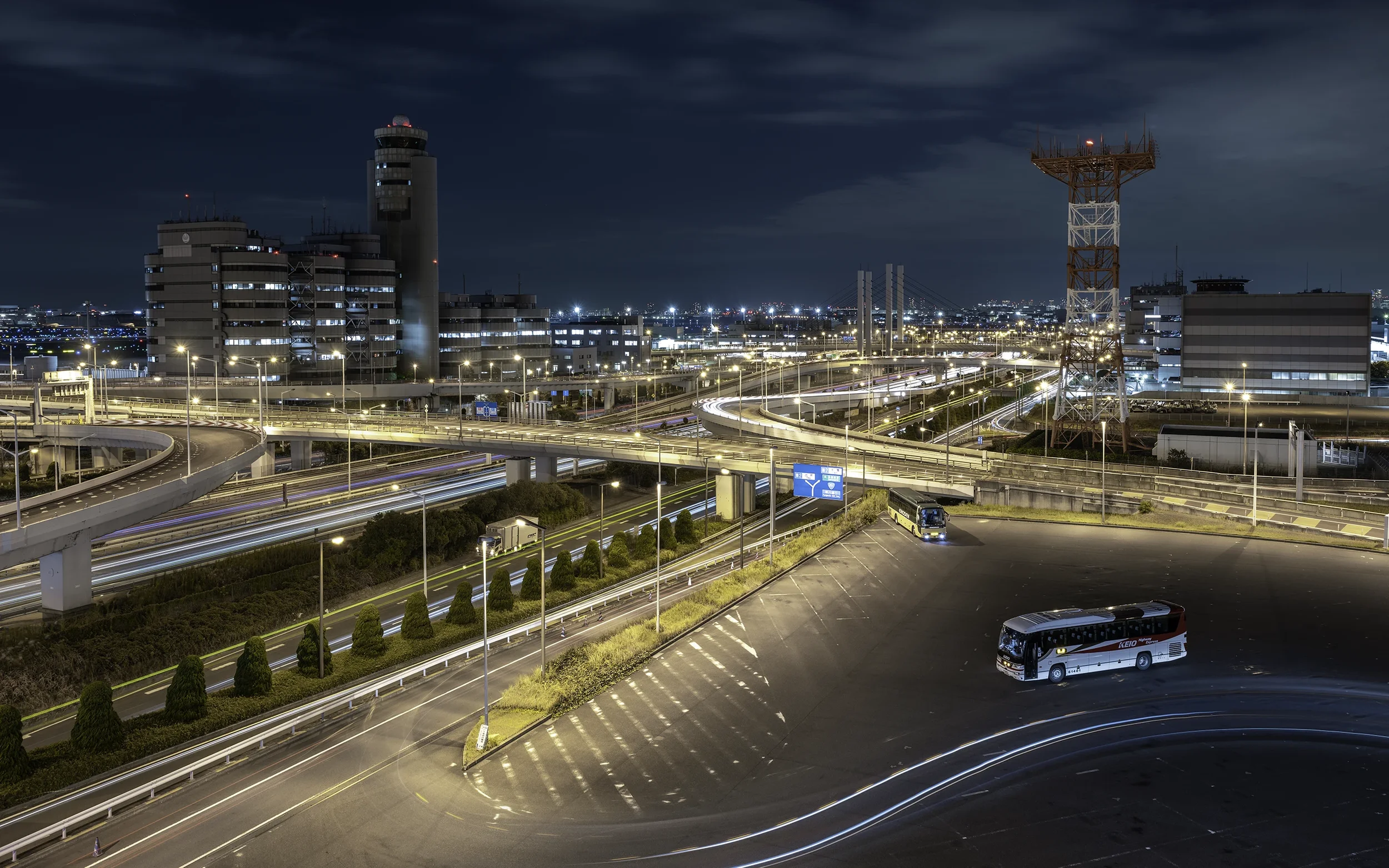 Vue nocturne de l'autoroute urbaine avec plusieurs voitures en mouvement, immeubles modernes et tours de communication dans la ville illuminée par des lampadaires.