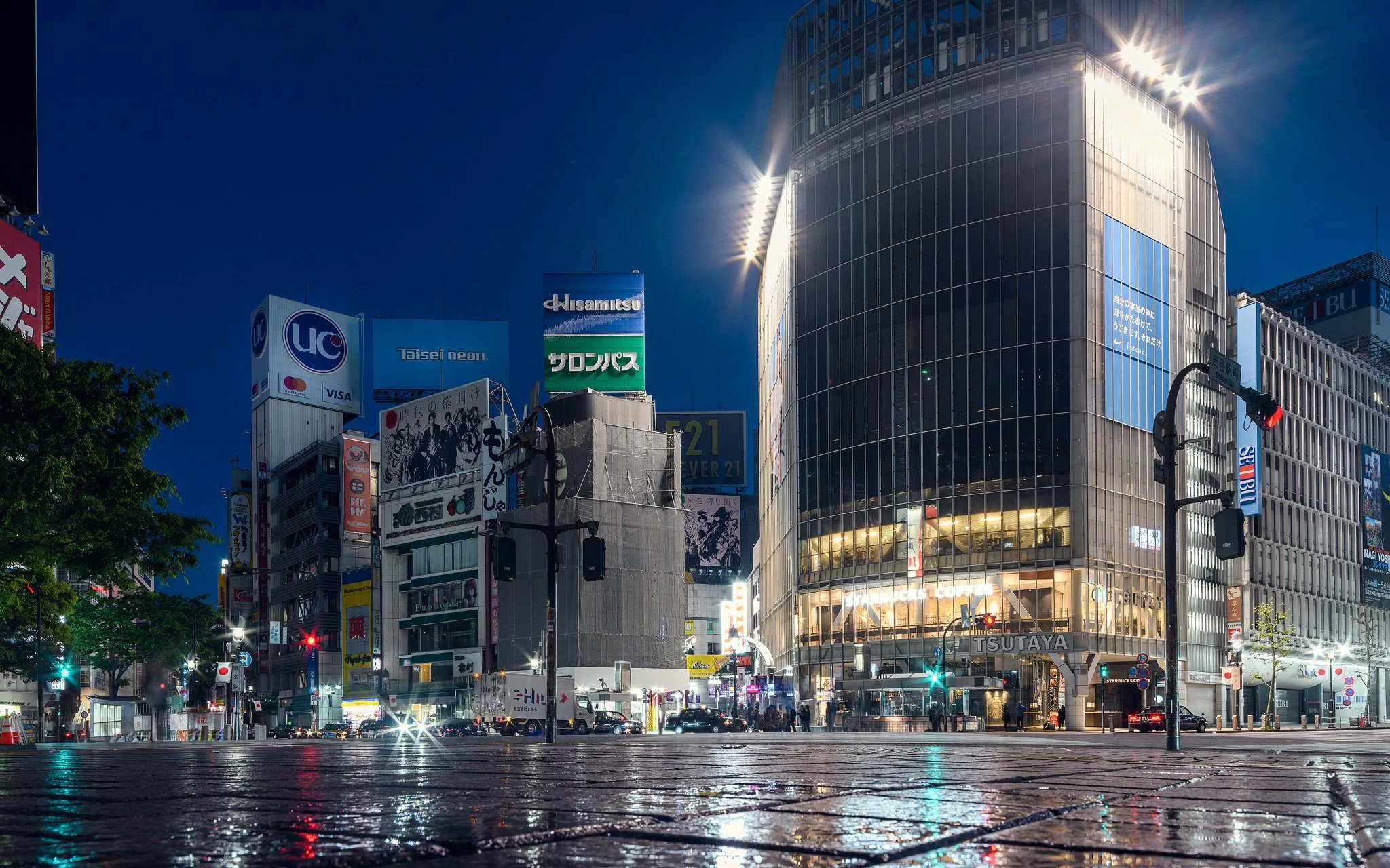 Une rue urbaine éclairée la nuit avec des bâtiments modernes en verre, des panneaux publicitaires en japonais, des feux de circulation et des passants.