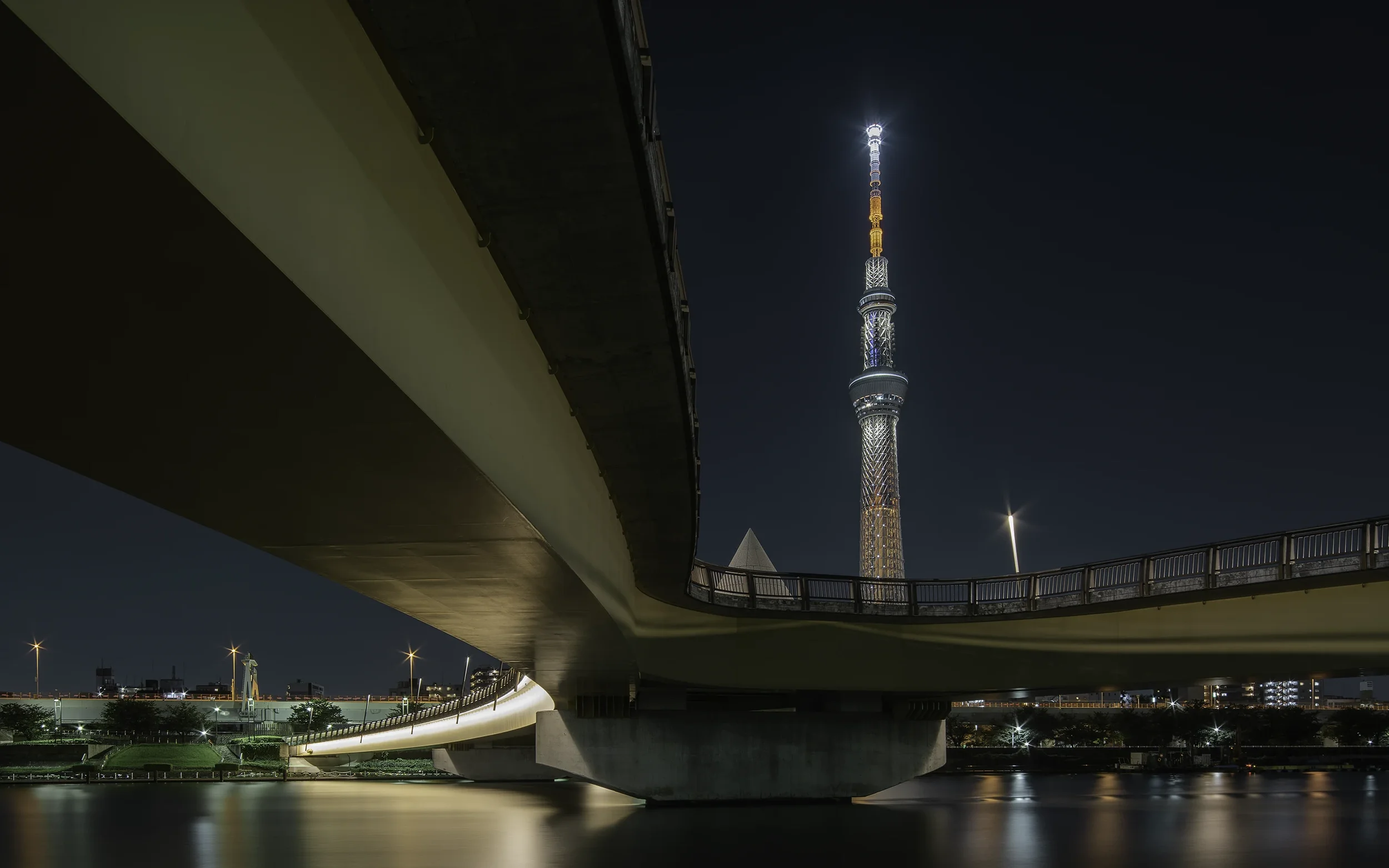 Vue nocturne d'un pont modern passant au-dessus d'une rivière, avec la tour de Tokyo illuminée en arrière-plan.