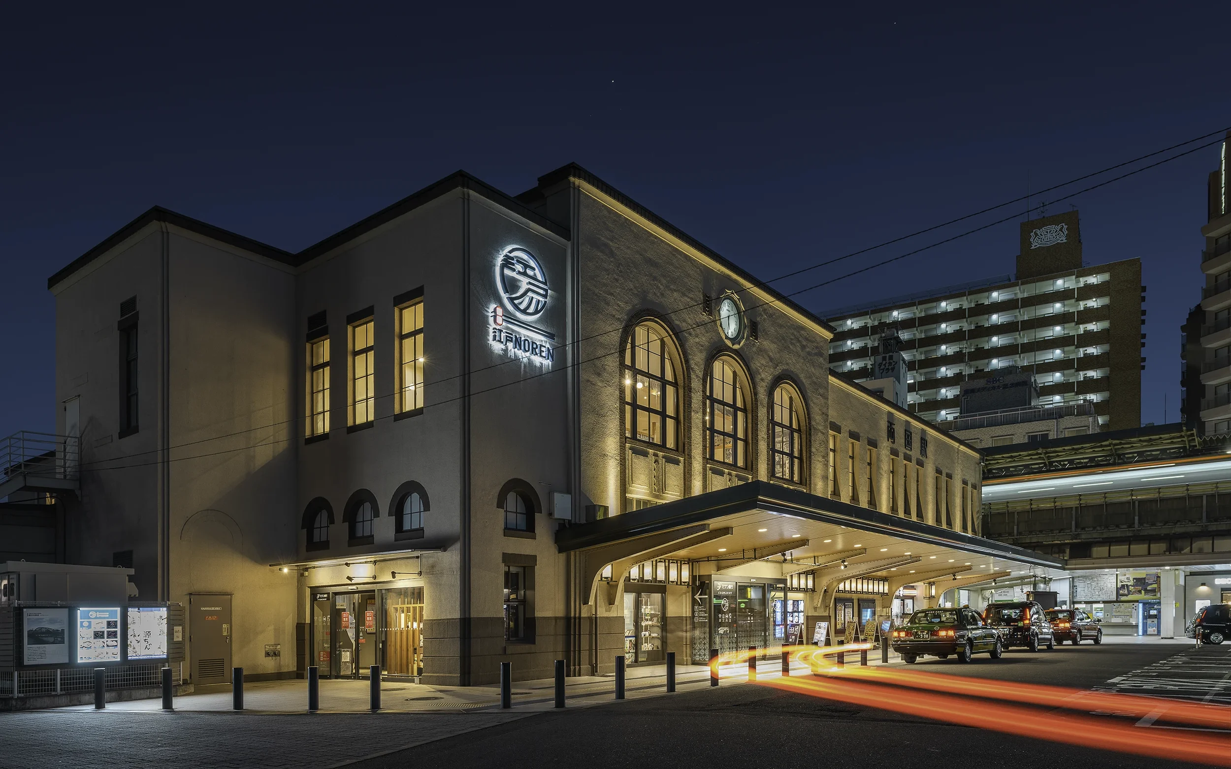 Bâtiment de style ancien avec des lumières chaudes, stationnement avec plusieurs voitures, éclairage nocturne et un logo lumineux en japonais sur la façade.