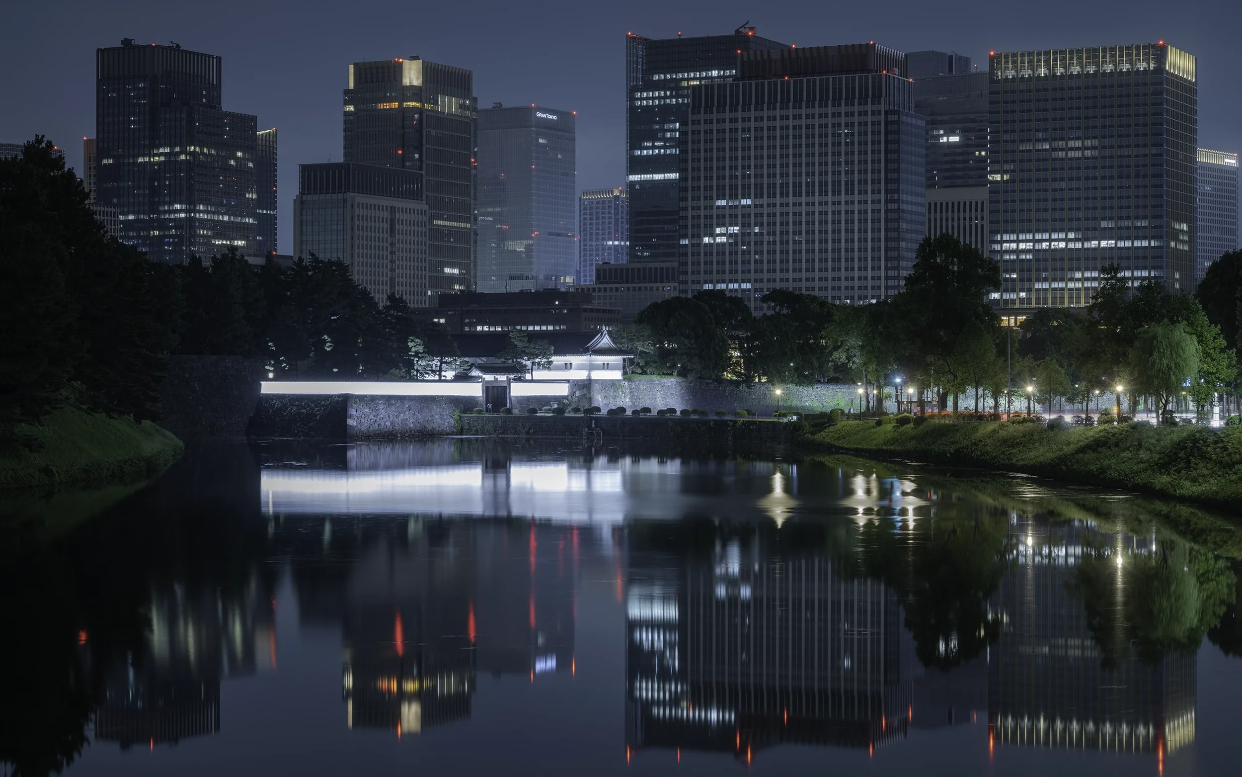Vue nocturne d'une skyline de gratte-ciel modernes illuminés, reflet dans une rivière calme, avec un parc arboré en premier plan.