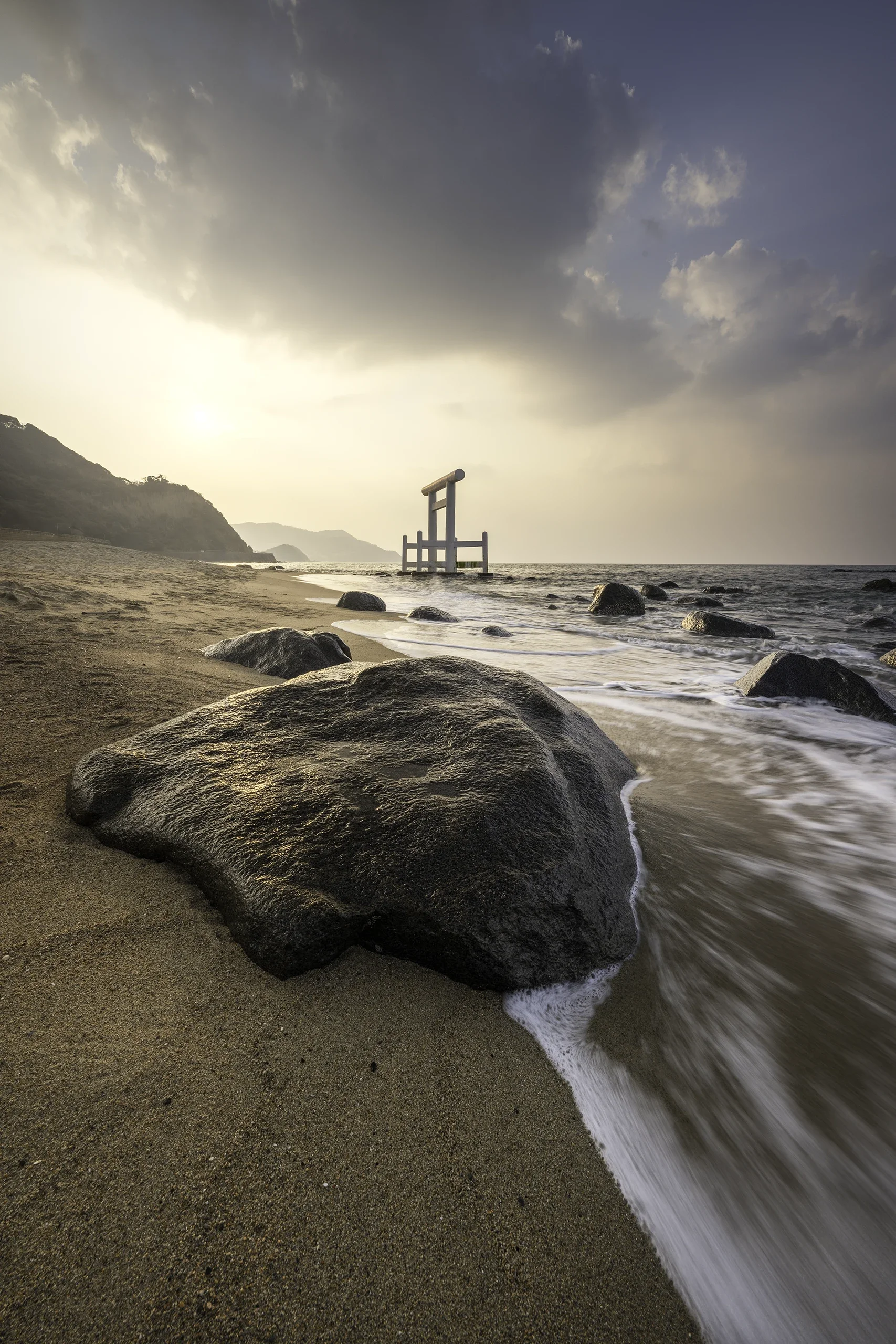 Plage avec des rochers, vague qui se retire, torii japonais en mer, montagnes au fond, ciel nuageux au coucher du soleil.