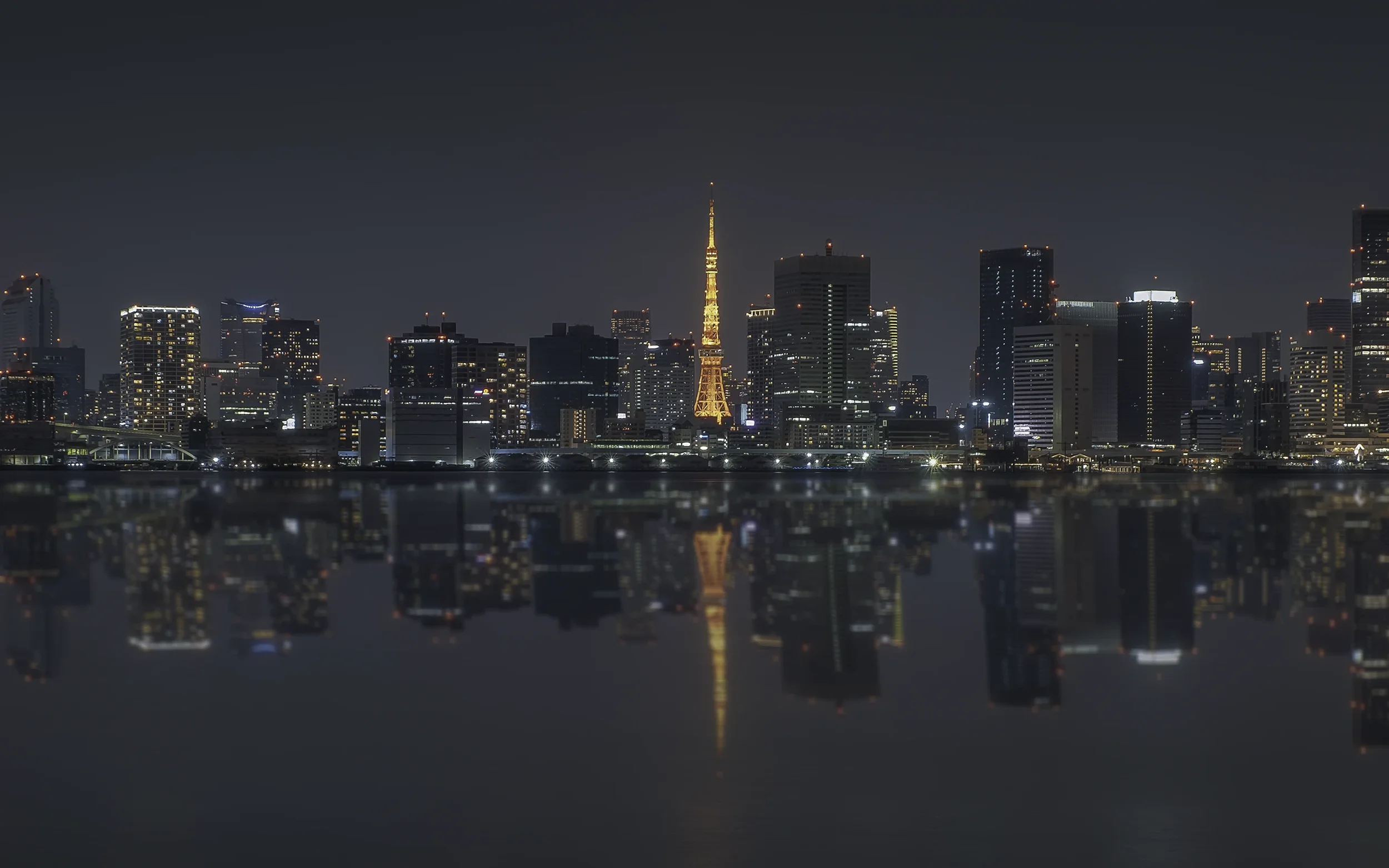 Vue de la ville de Tokyo la nuit avec la tour de Tokyo illuminée en orange et son reflet dans l'eau devant un ciel sombre.