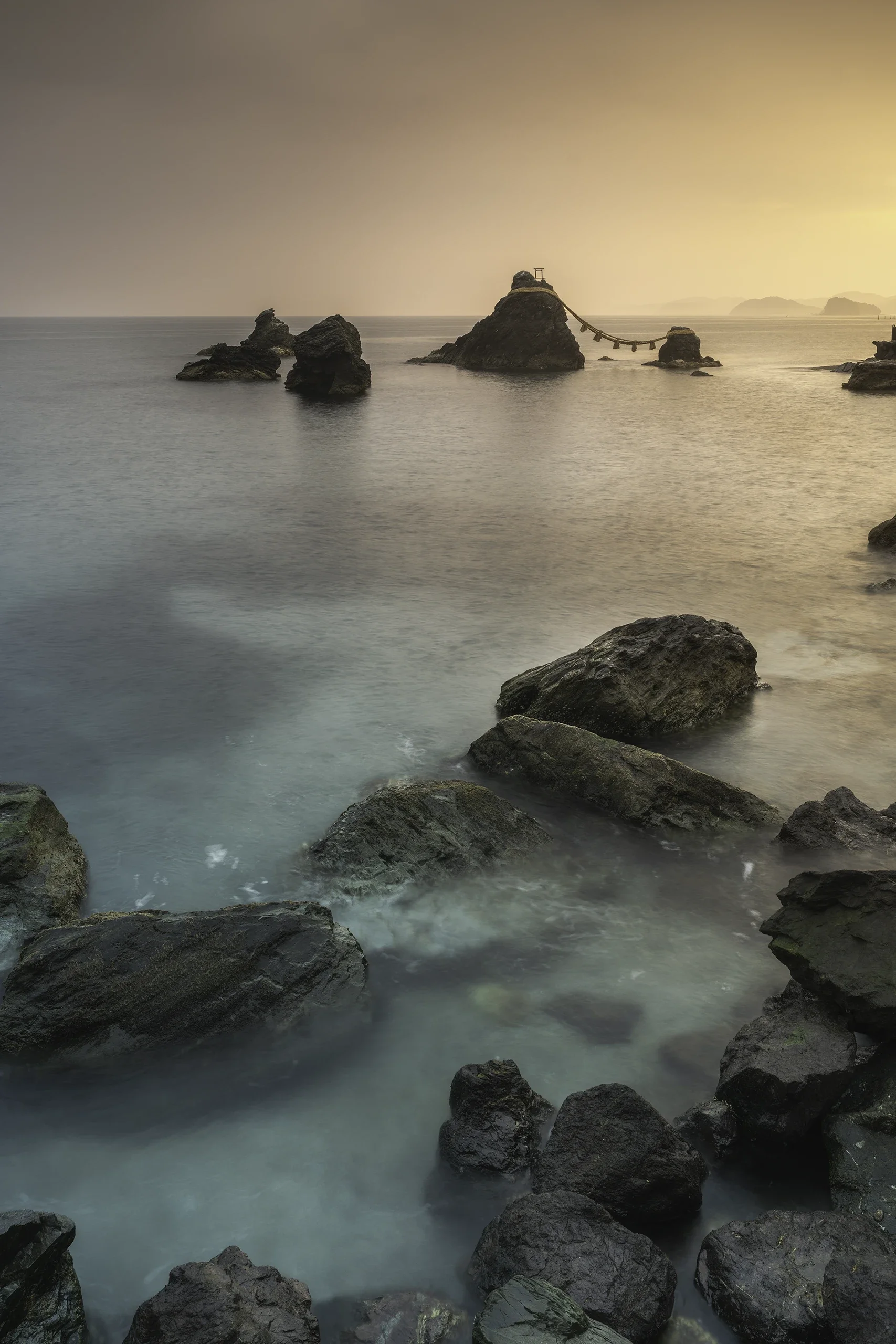Scène de plage au coucher du soleil avec des rochers dans la mer et une structure en forme de torii flottante dans l'eau au loin.