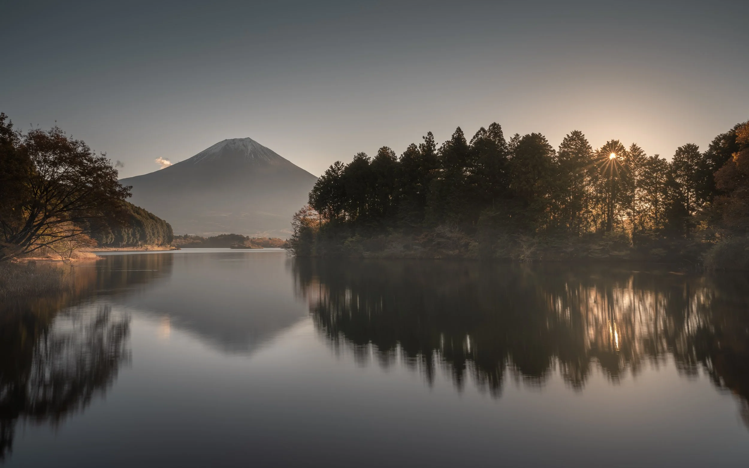 Paysage avec un lac calme, des arbres au bord de l'eau, un mont en arrière-plan, et le soleil couchant derrière les arbres.
