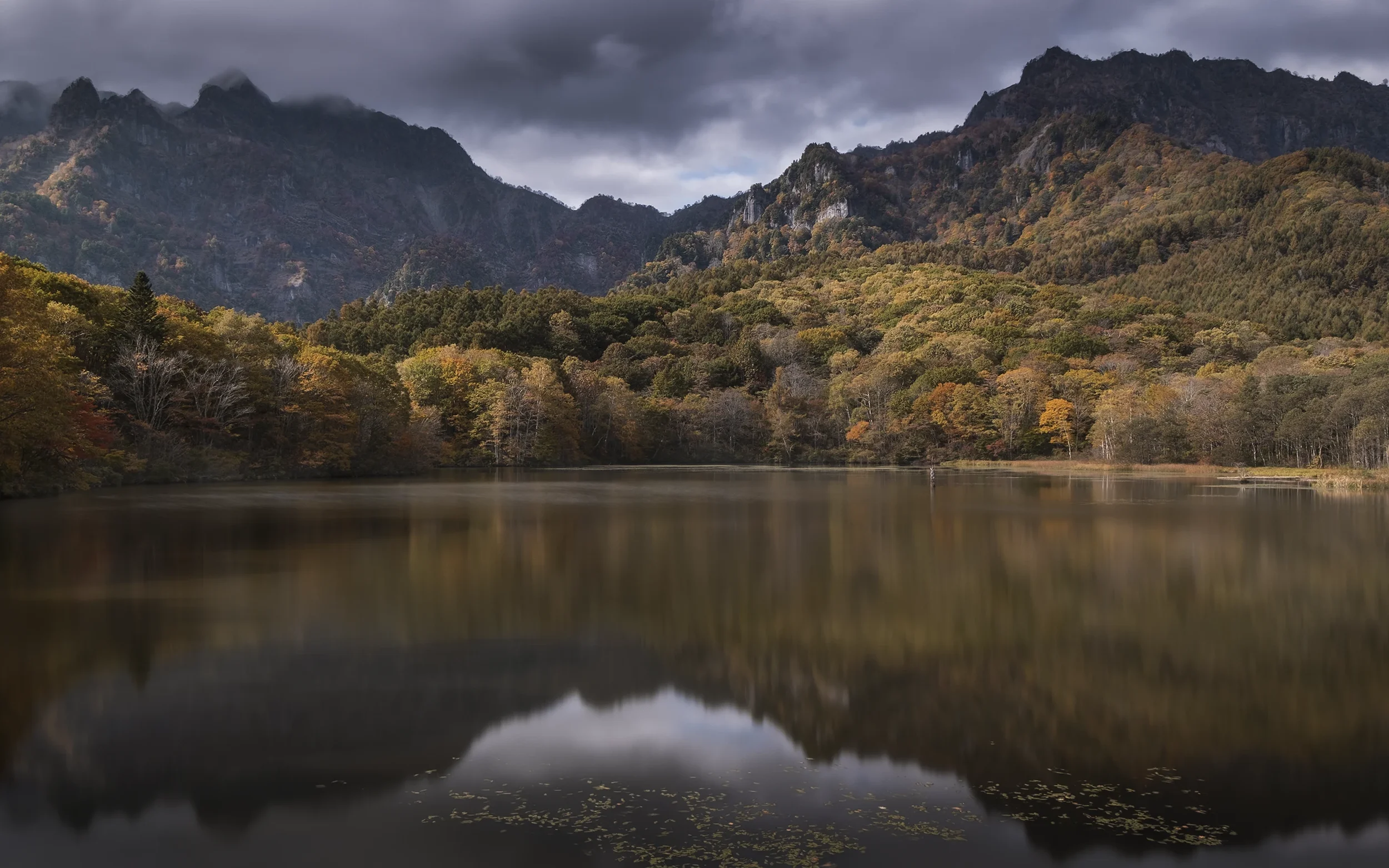 Scène de nature avec un lac reflétant la forêt colorée d'automne et les montagnes sous un ciel nuageux.