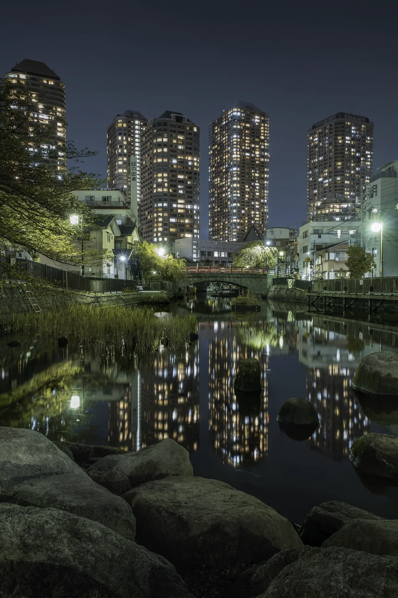 Une vue nocturne d'une rivière avec des pierres au premier plan, des bâtiments résidentiels et commerciaux illuminés en arrière-plan, et un reflet de la ville dans l'eau.