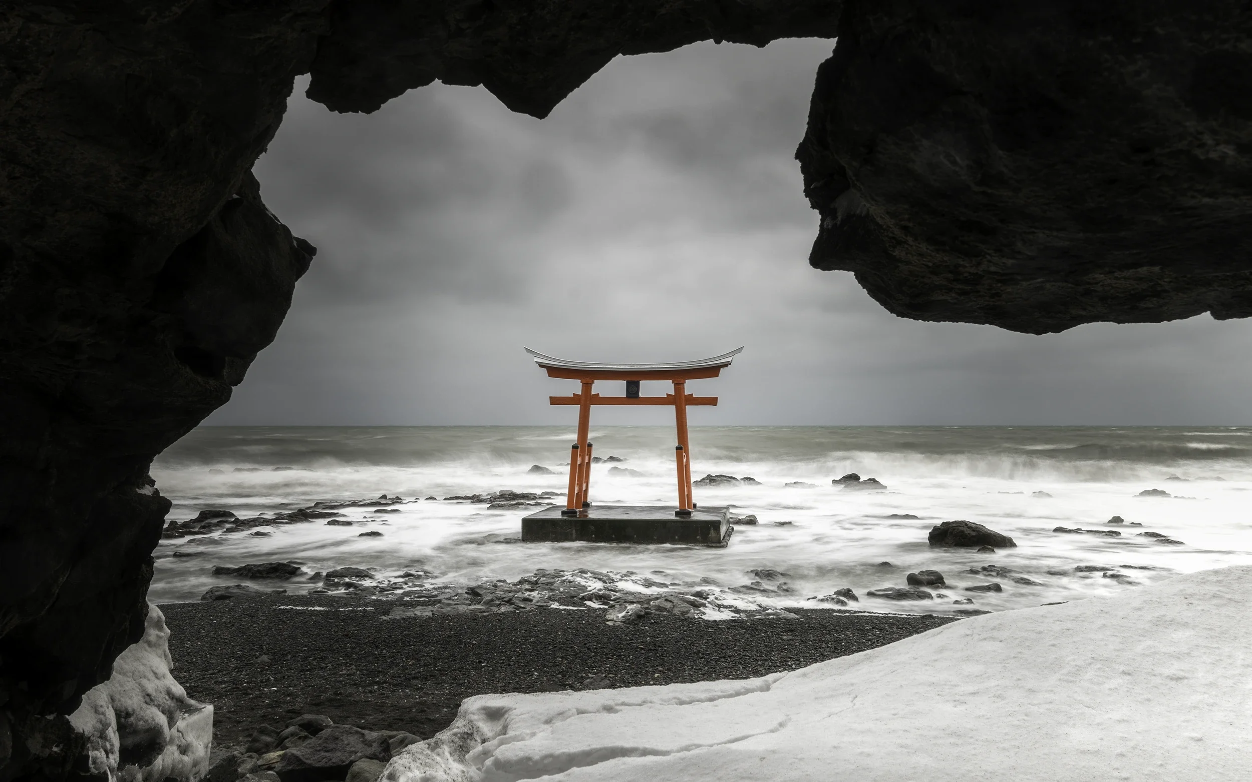 Vermilion torii gate standing in the sea during winter on the coast of Hokkaido, framed by snow and dramatic storm clouds.