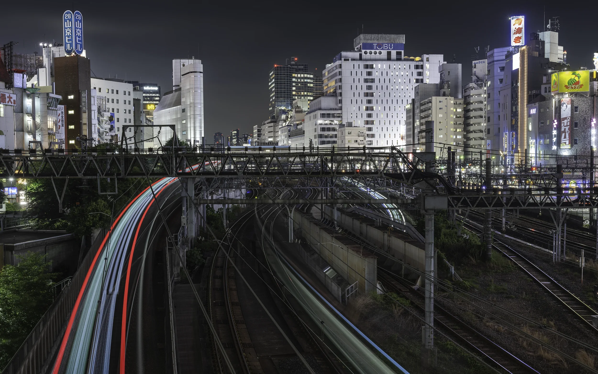 Ville de nuit avec des bâtiments lumineux et des voies de train éclairées, montrant des trains en mouvement à cause des trains lumineux et du mouvement long.