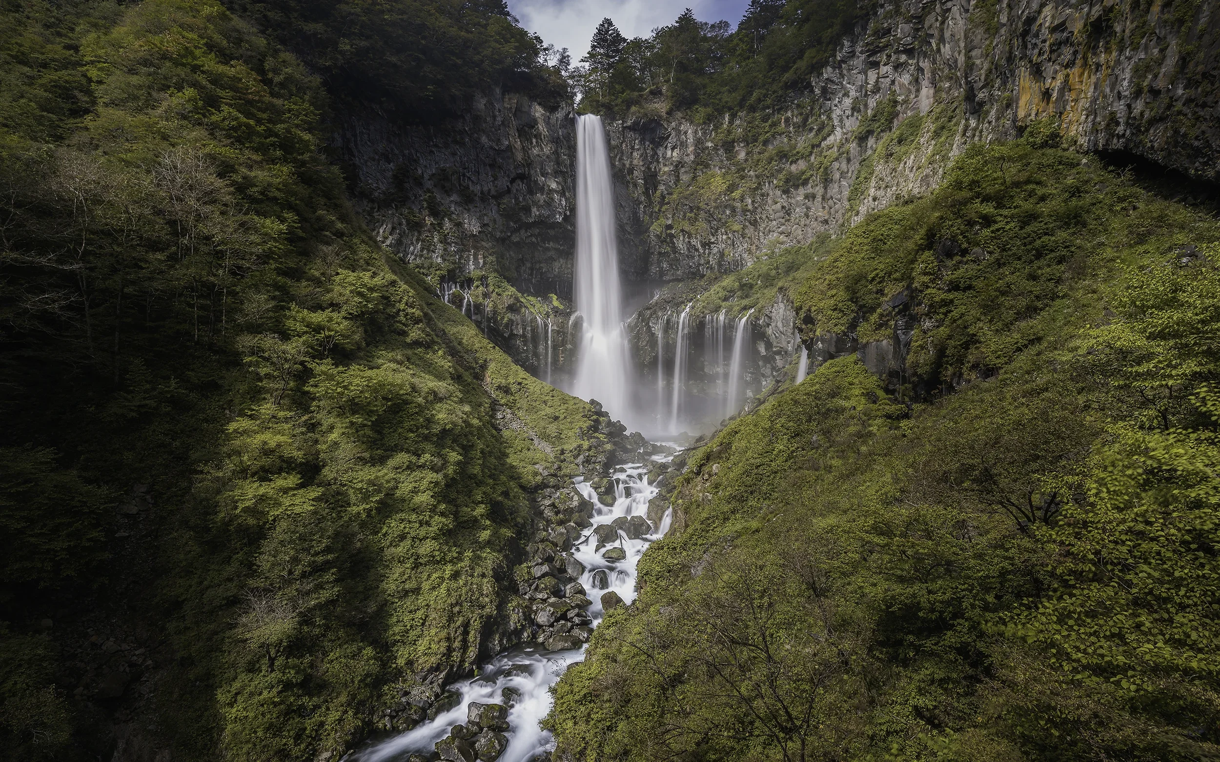 Cascade d'eau tombant d'une falaise dans une vallée verdoyante avec des arbres et des rochers.