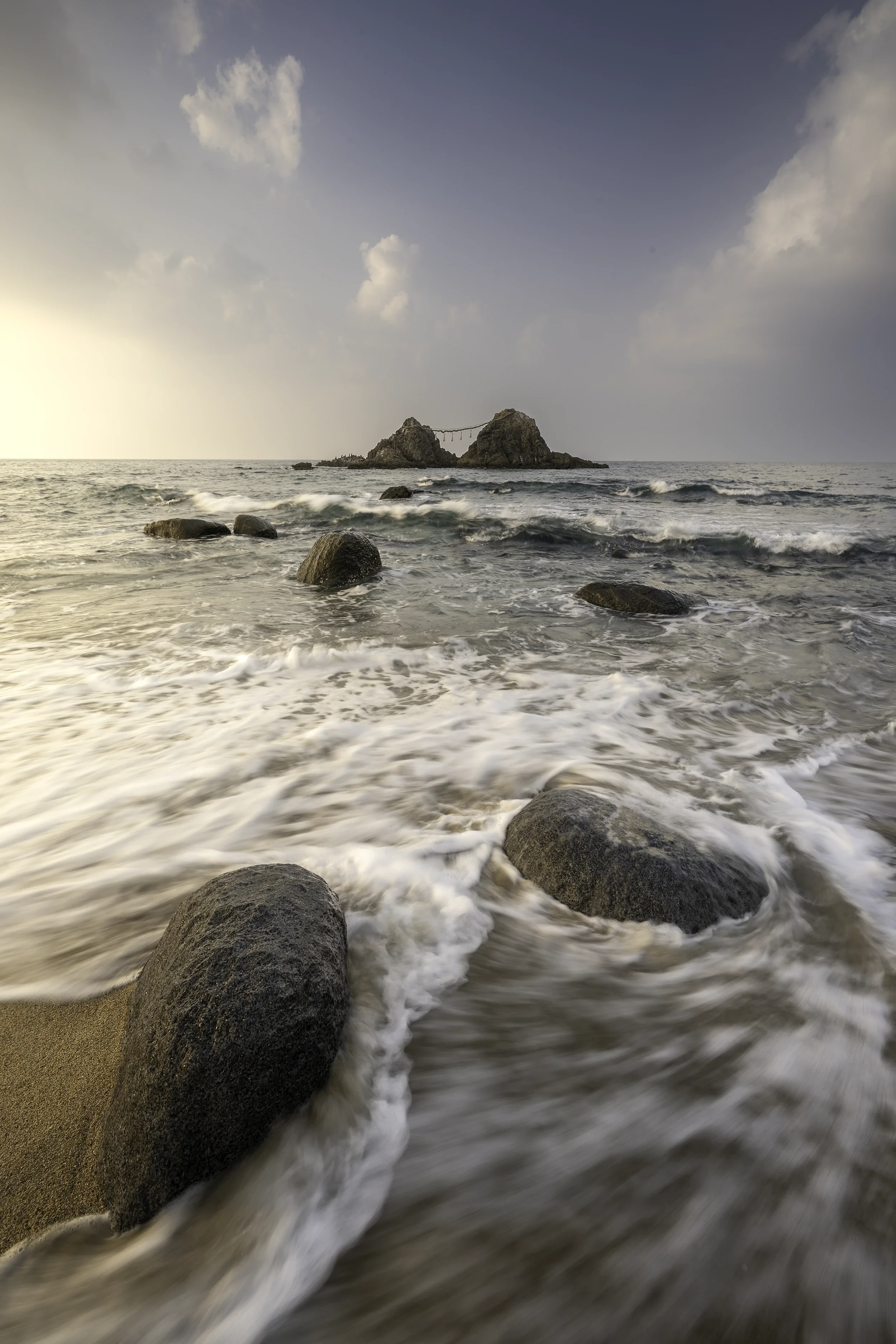 The famous Meoto Iwa of Kyushu photographed at sunset, where sacred couple rocks emerge from the sea beneath soft golden light and rolling ocean waves.