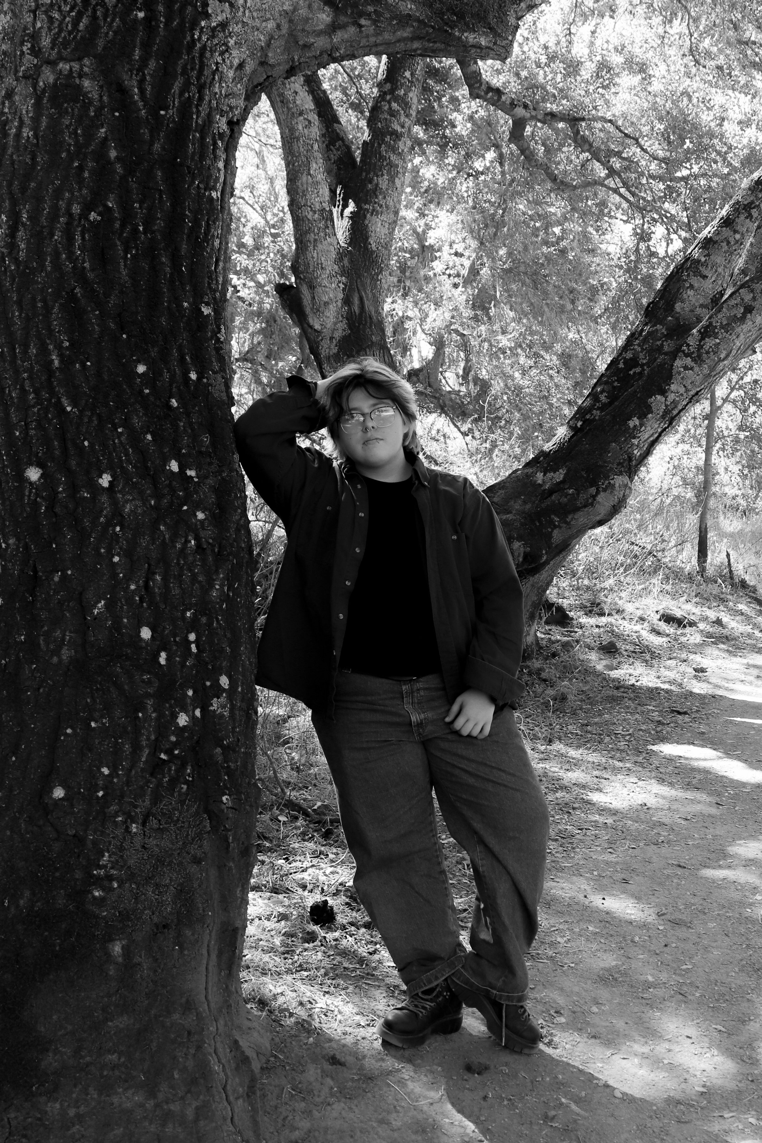 A person with glasses and medium-length hair leaning against a large tree trunk in a natural outdoor setting, with sunlight filtering through the leaves.