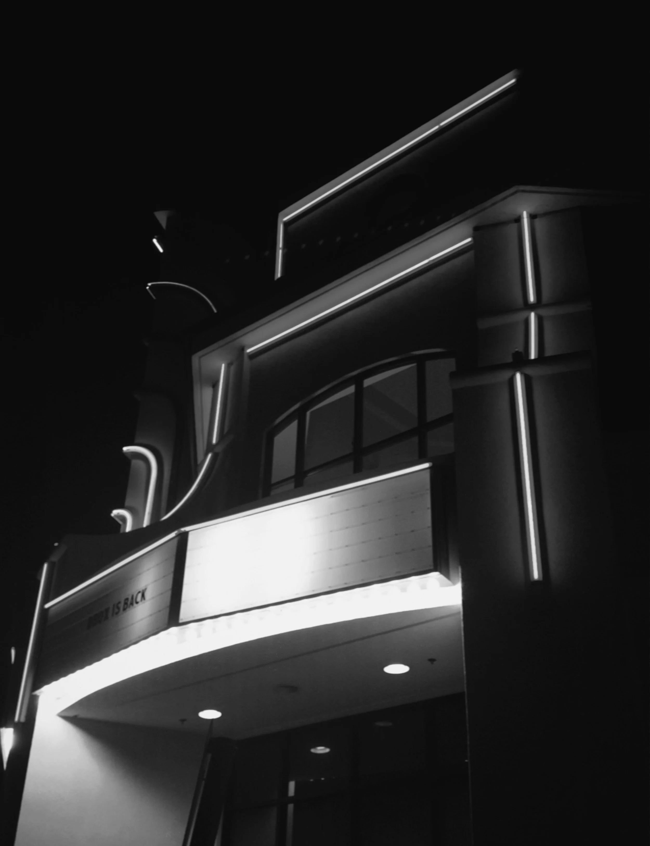 A neon-lit building facade at night, featuring signage and architectural details, with the words "THEATER IS BACK" visible on a sign.