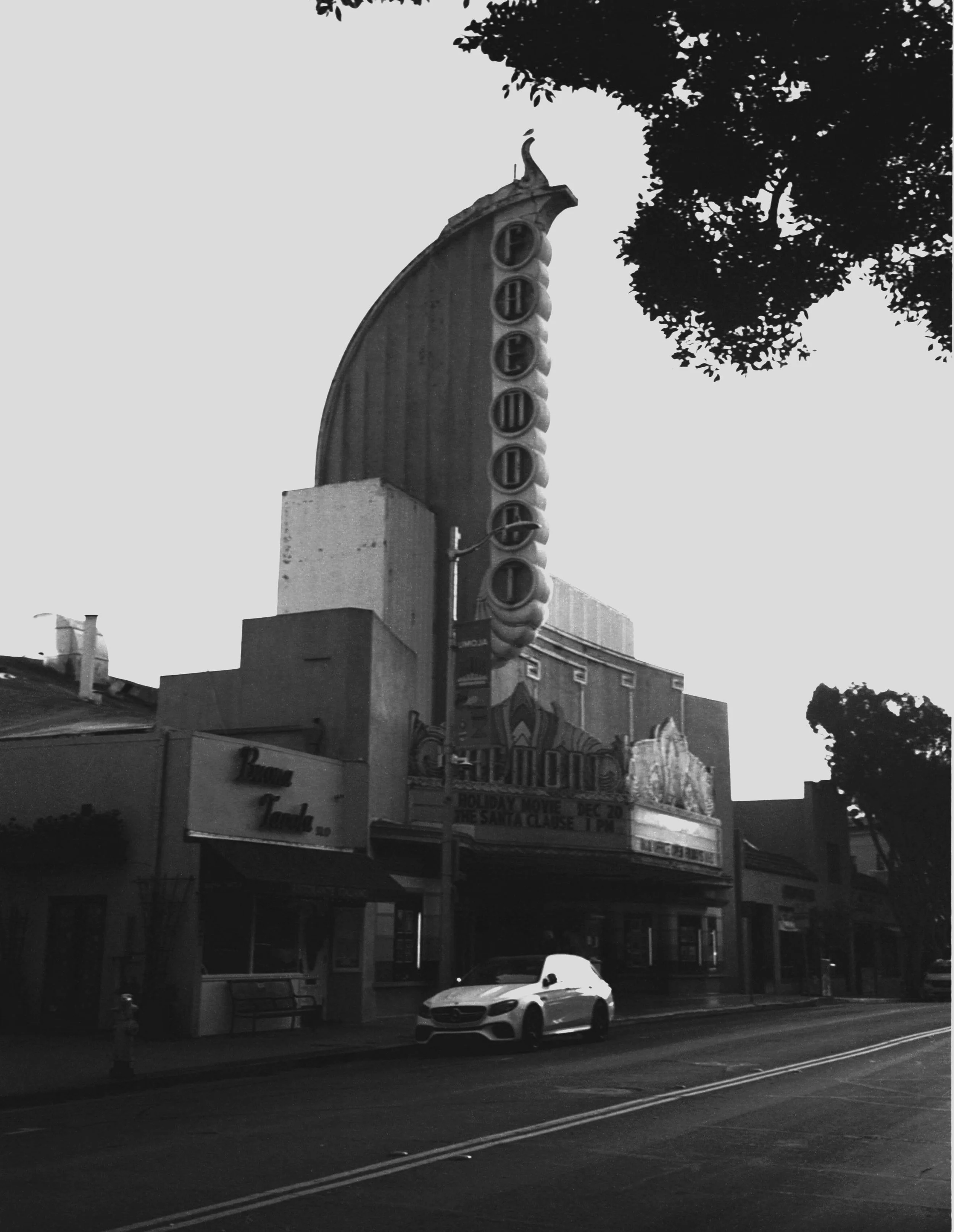 Historic movie theater with a tall, vertical sign displaying the word 'FAIRMONT' and a marquee advertising a holiday movie event, with a modern car parked in front and trees partially shading the scene.