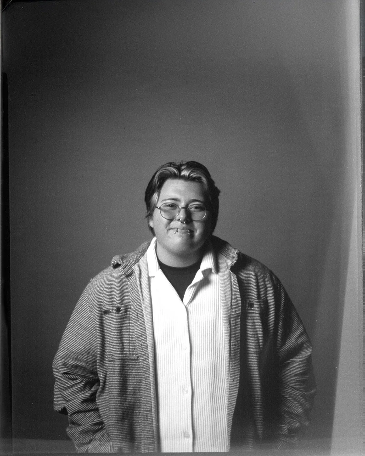 Black and white photo of a person with short hair, glasses, and piercings, wearing a striped shirt and a jacket, smiling at the camera.