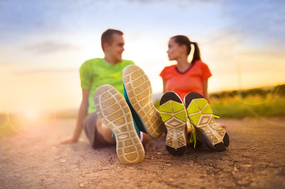 A man and woman sitting on the ground with their shoes in the foreground, smiling at each other during sunset.