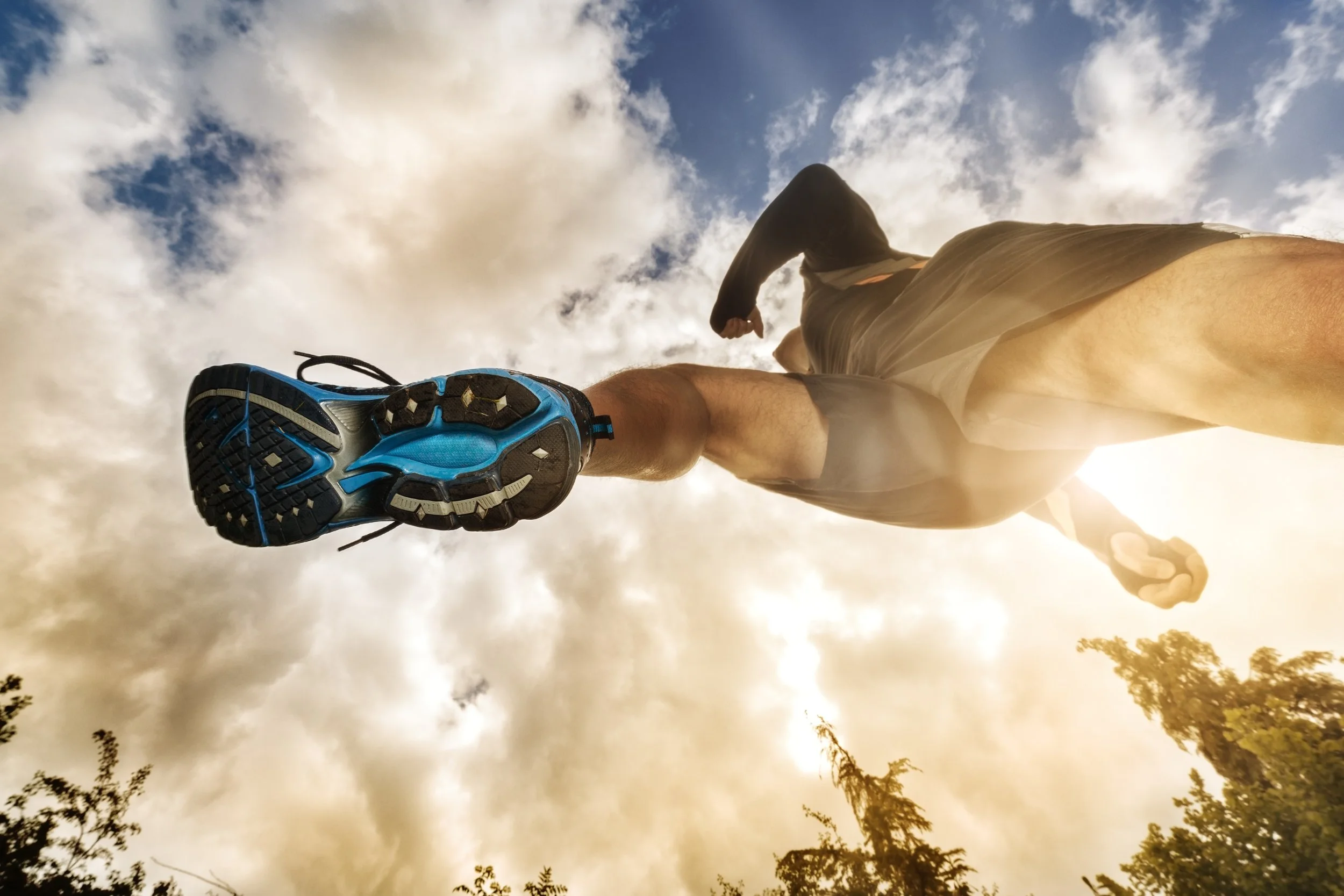 A person running outdoors with a cloudy sky background, viewed from a low angle, showing the bottom of their athletic shoe and part of their body.