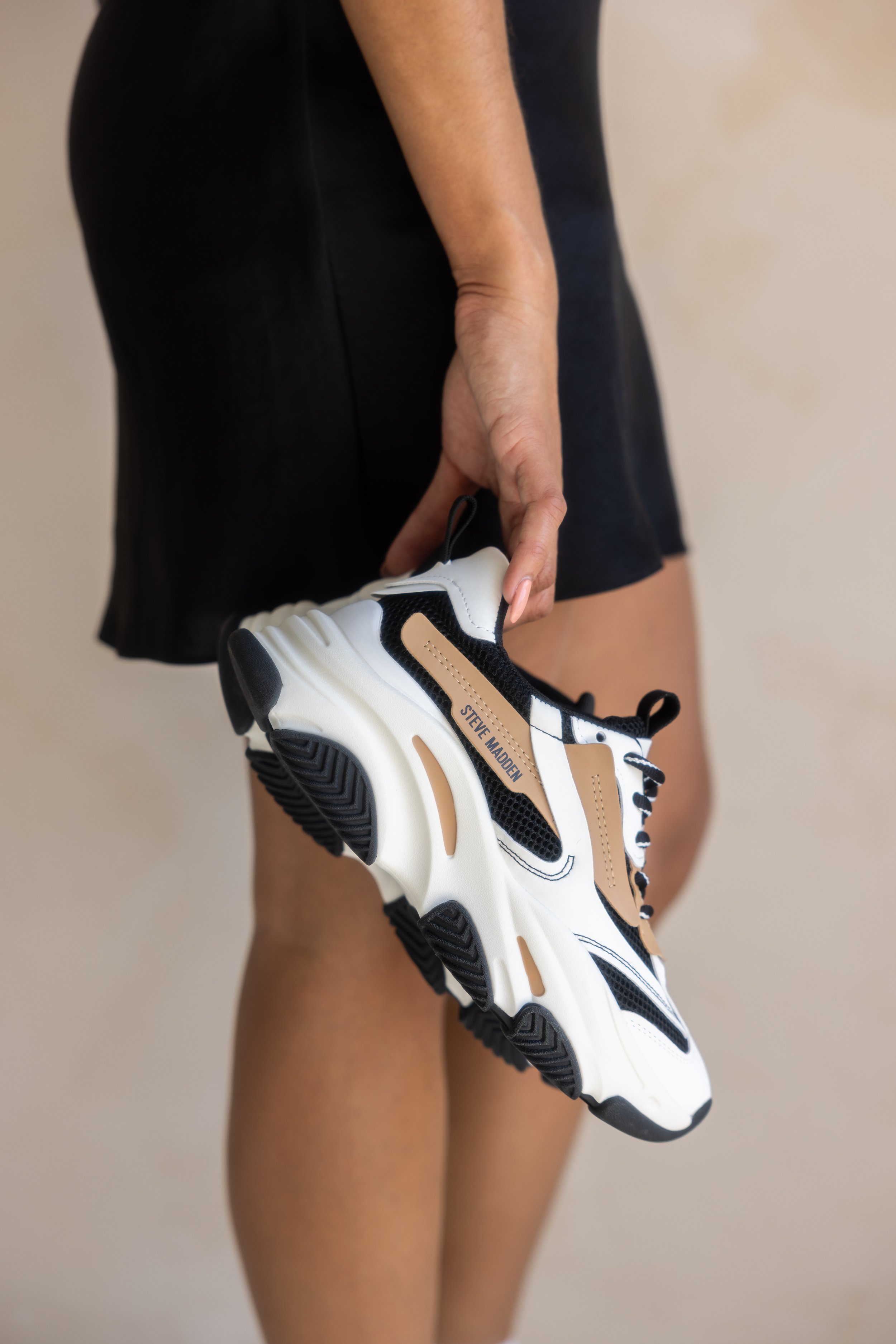 Person holding a white, black, and beige sneaker with the brand 'Steve Madden' visible on the side.