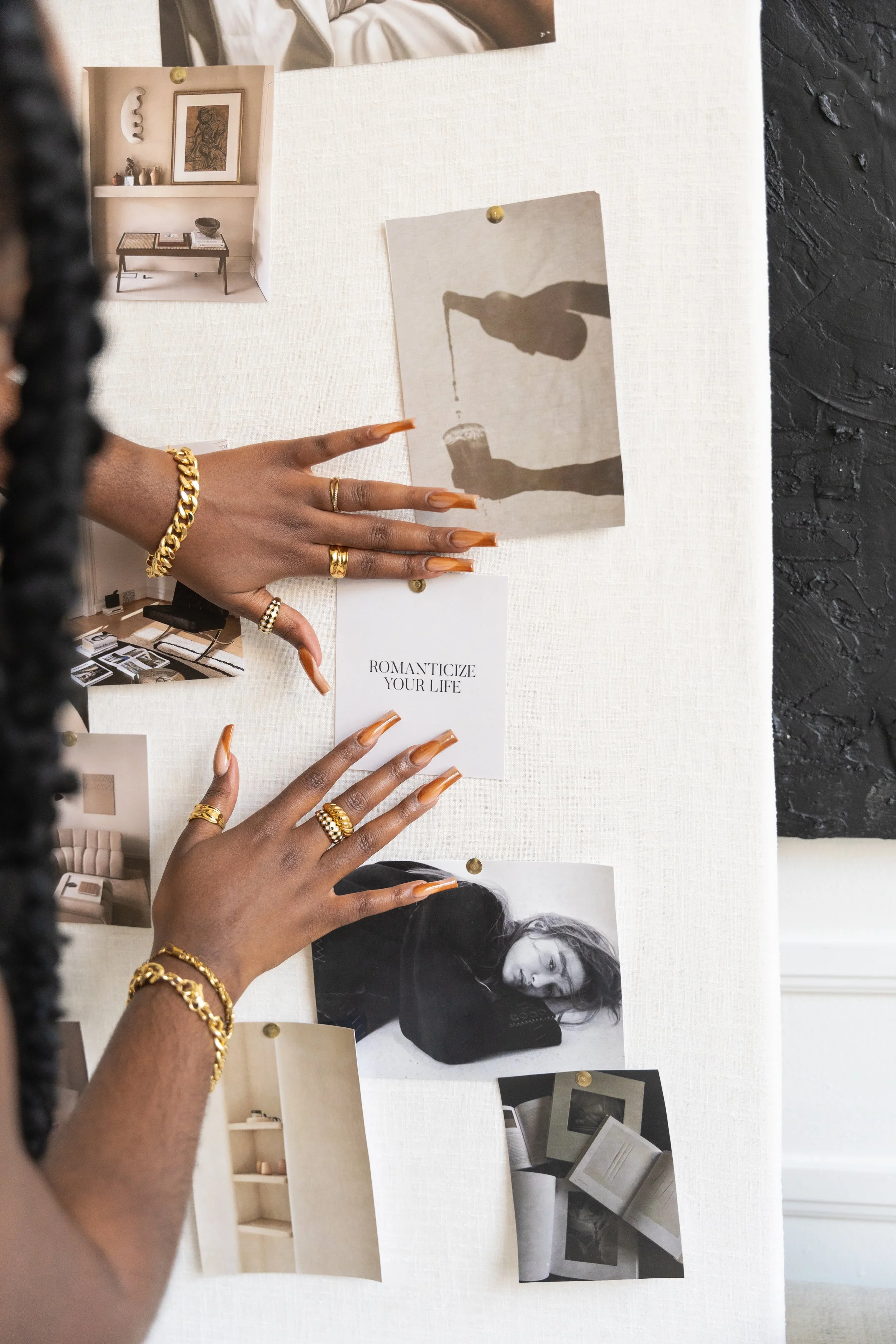 Person with gold jewelry arranging photos and notes on a white corkboard, including a monochrome photo of a woman with her head resting on her arm, and a printed quote 'ROMANTICIZE YOUR LIFE'.