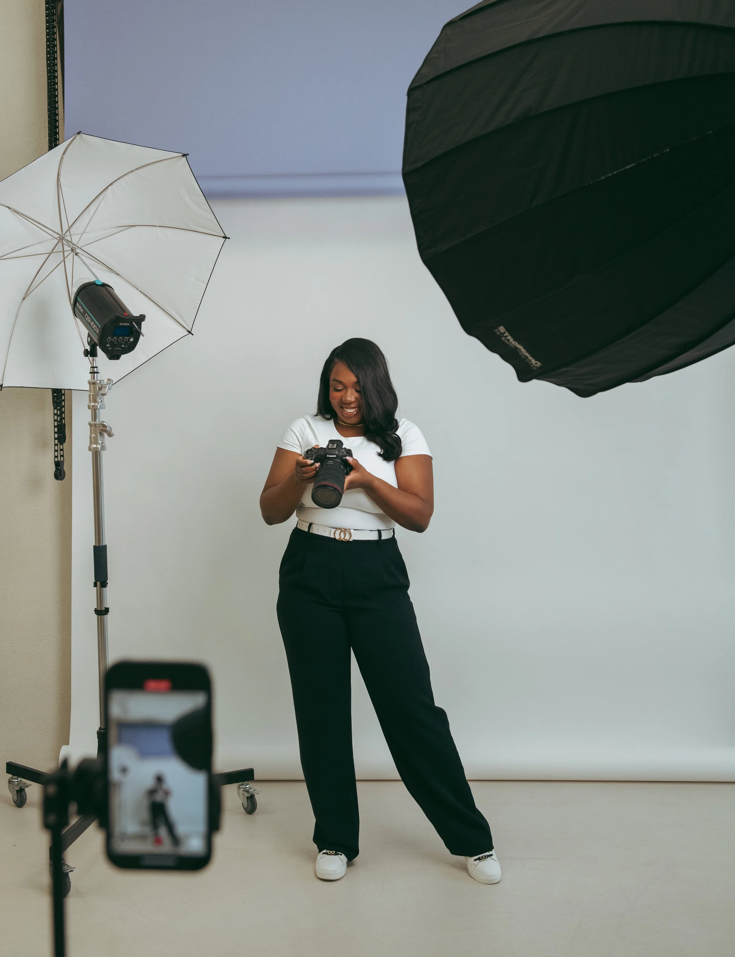 A woman in a photo studio holding a camera, smiling, with photography lighting equipment and a white backdrop.
