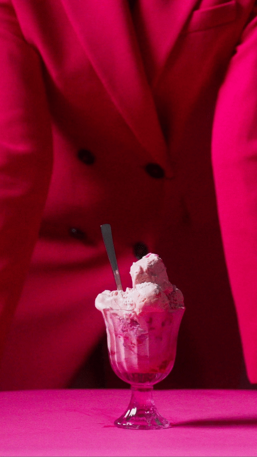 A person in a pink blazer standing behind a glass filled with ice cream, with a spoon in the ice cream.