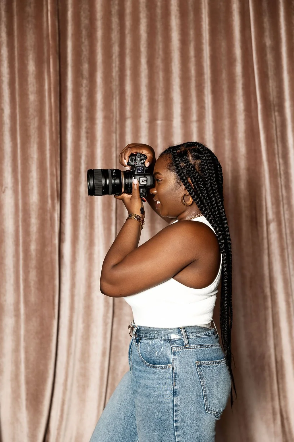 Young woman with braided hair and hoop earrings taking a photo with a professional camera against a brown curtain backdrop.