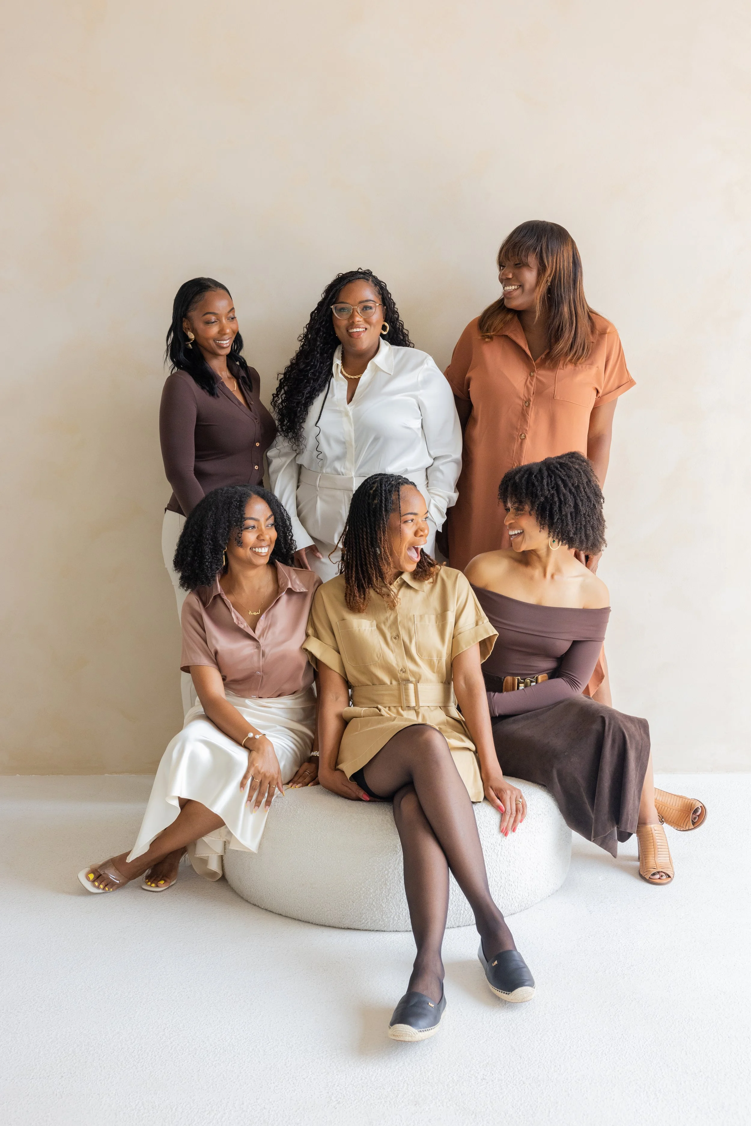 Group of seven women smiling and laughing together against a plain beige wall, some sitting on a circular white platform and others standing behind.