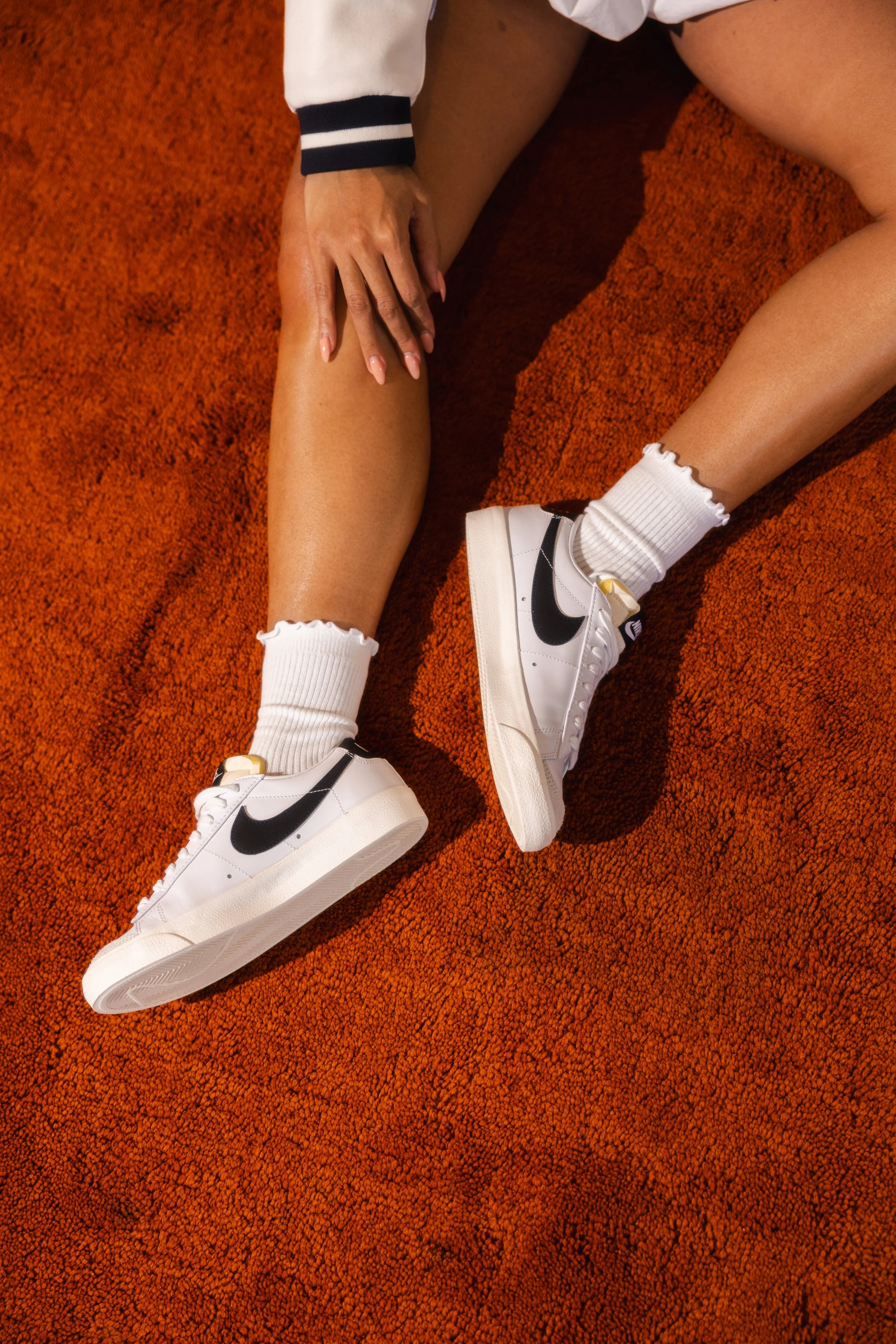 A person is sitting on an orange textured carpet, wearing white Nike sneakers with black Nike swoosh logos, white ankle socks with ruffled edges, and a white jacket with black stripes on the sleeves. They are touching their left leg with their left hand.