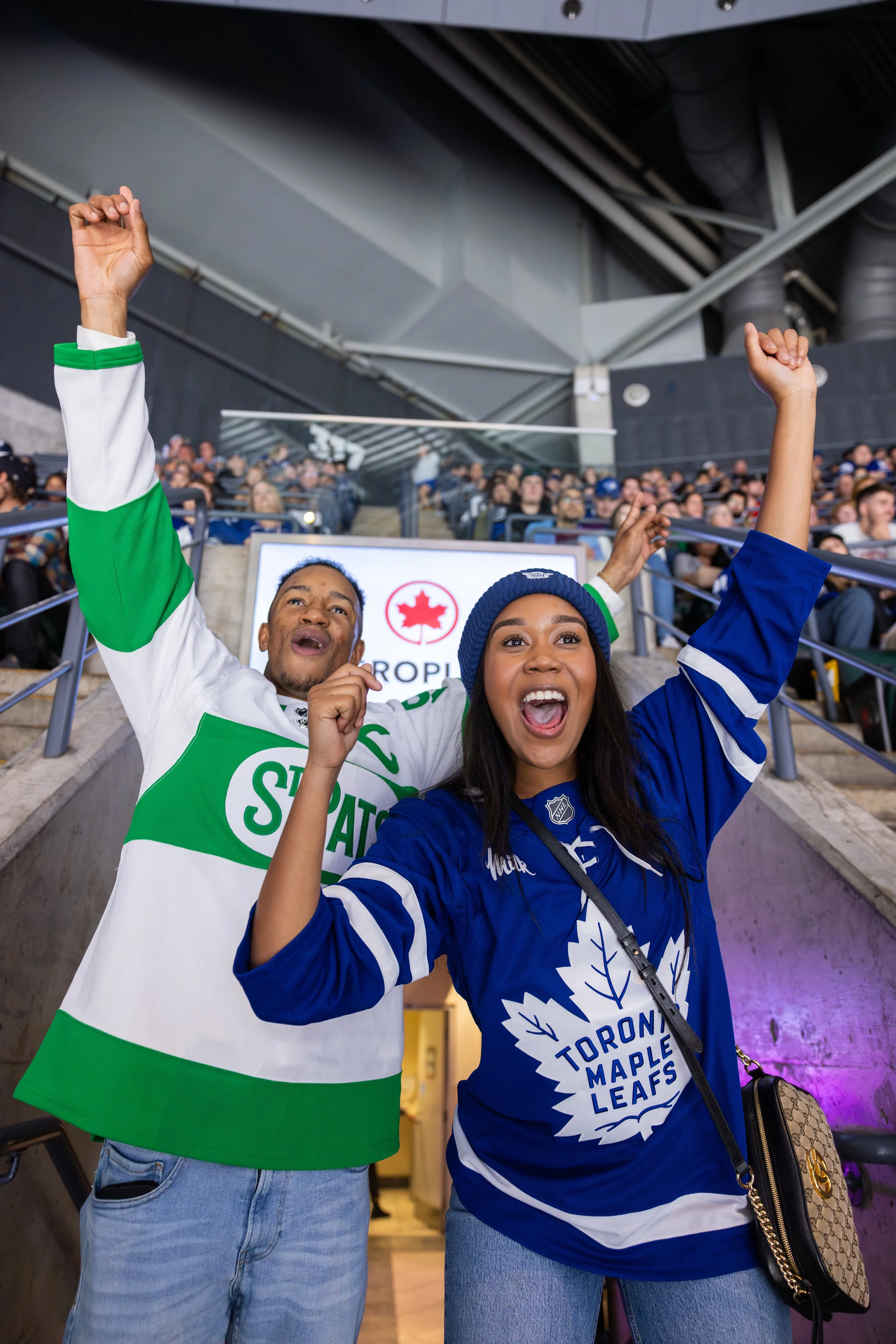 Two hockey fans celebrating, wearing Toronto Maple Leafs and Sporting jerseys, with people in the background at a stadium.