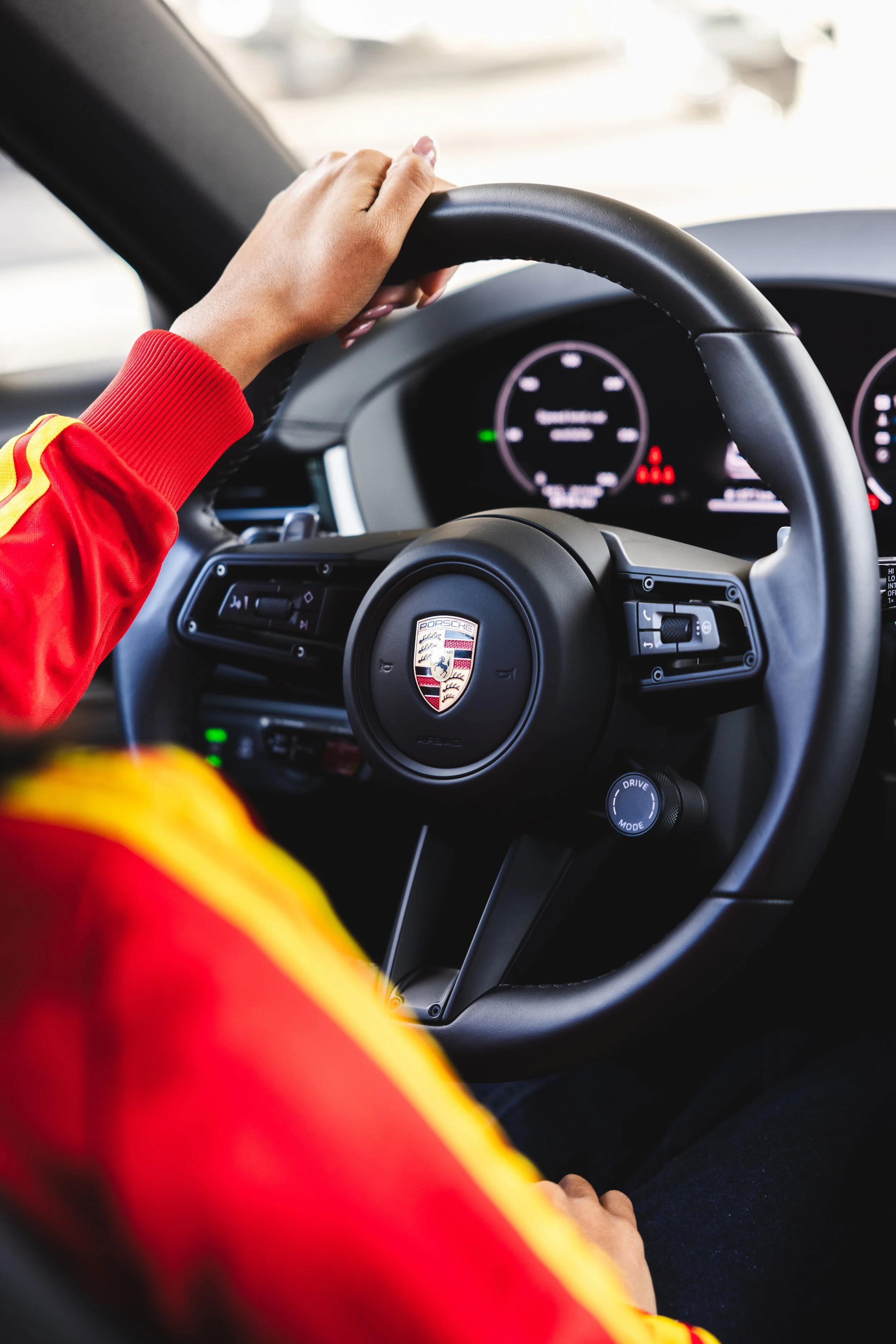 Close-up of a person with a red and yellow jacket holding a Porsche steering wheel inside a car.