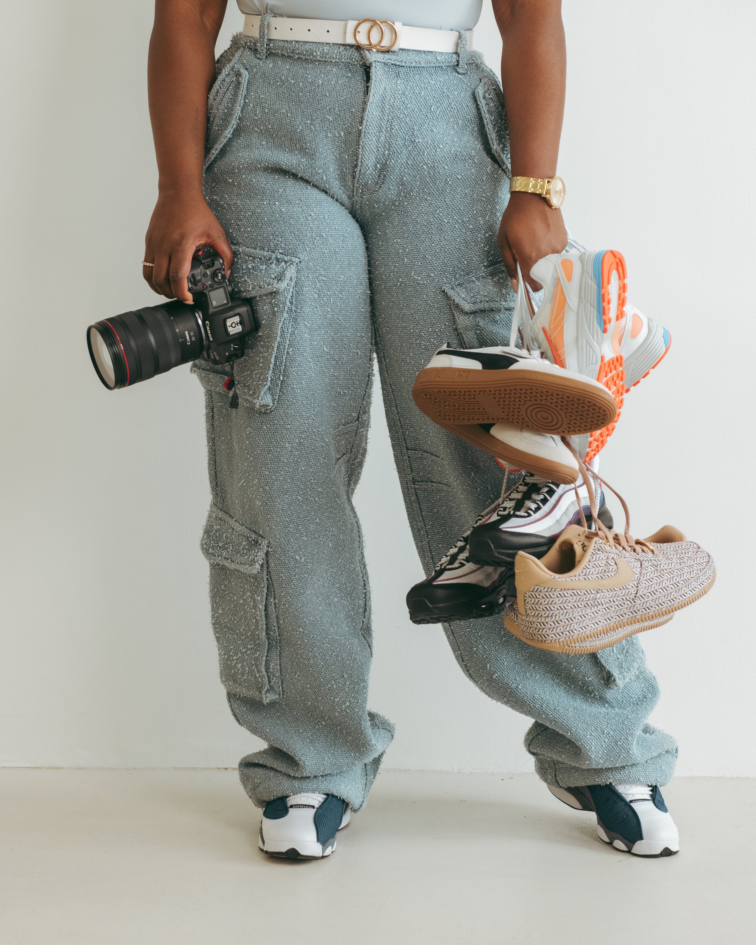 Person wearing light blue cargo pants with a white belt, holding a camera in one hand and a bunch of sneakers in the other, including brand names like Nike and Puma, standing against a plain background.