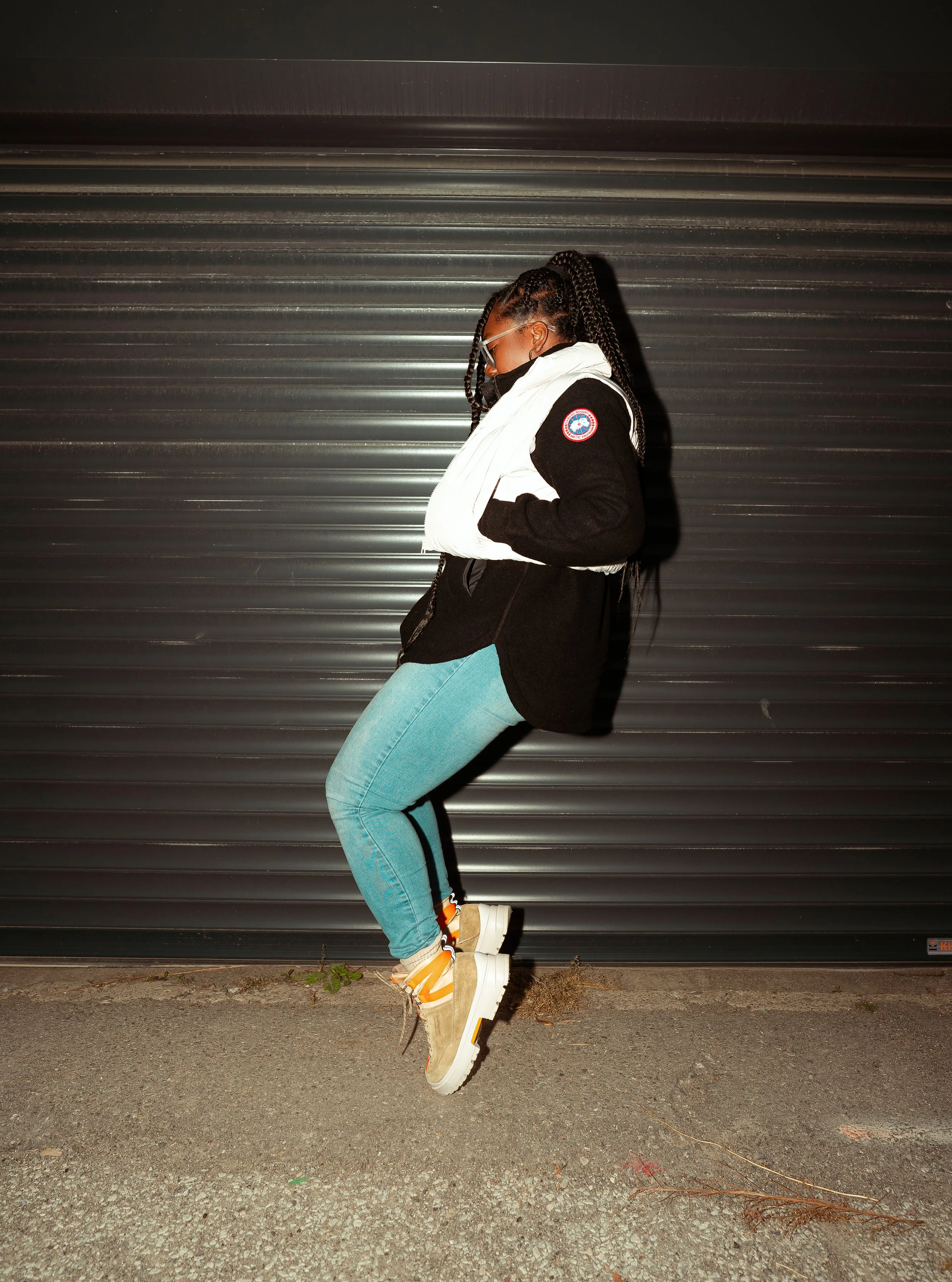 A person jumping in front of a metal shutter door at night, wearing a black hoodie, white vest, blue jeans, tan boots, sunglasses, and a face mask.