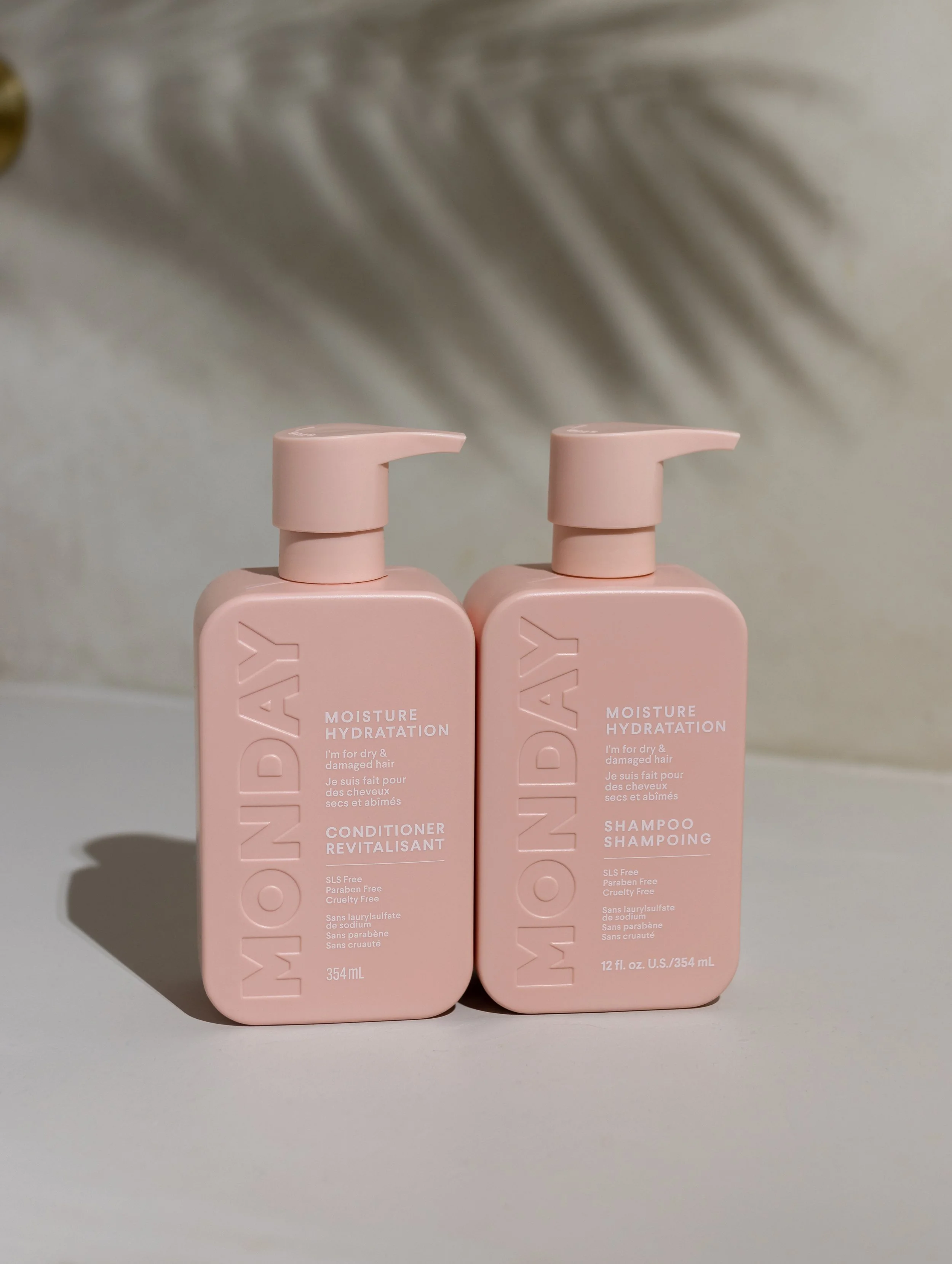 Two pink bottles of MONDAY hair products, Moisture Hydration Conditioner and Shampoo, with pump dispensers, on a neutral surface with a blurred background.