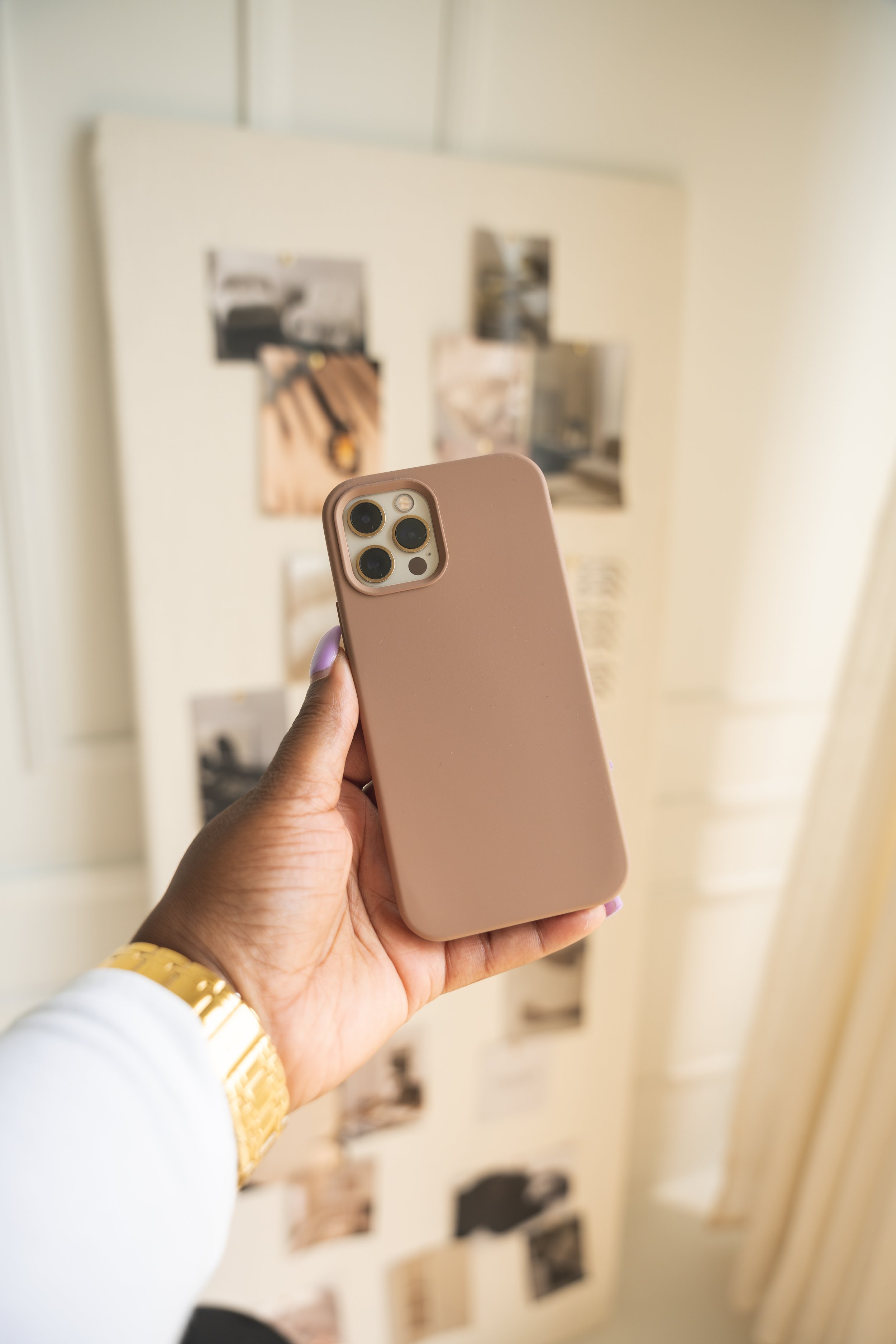 A hand holding a pink smartphone in front of a beige wall with photos and notes pinned on it.