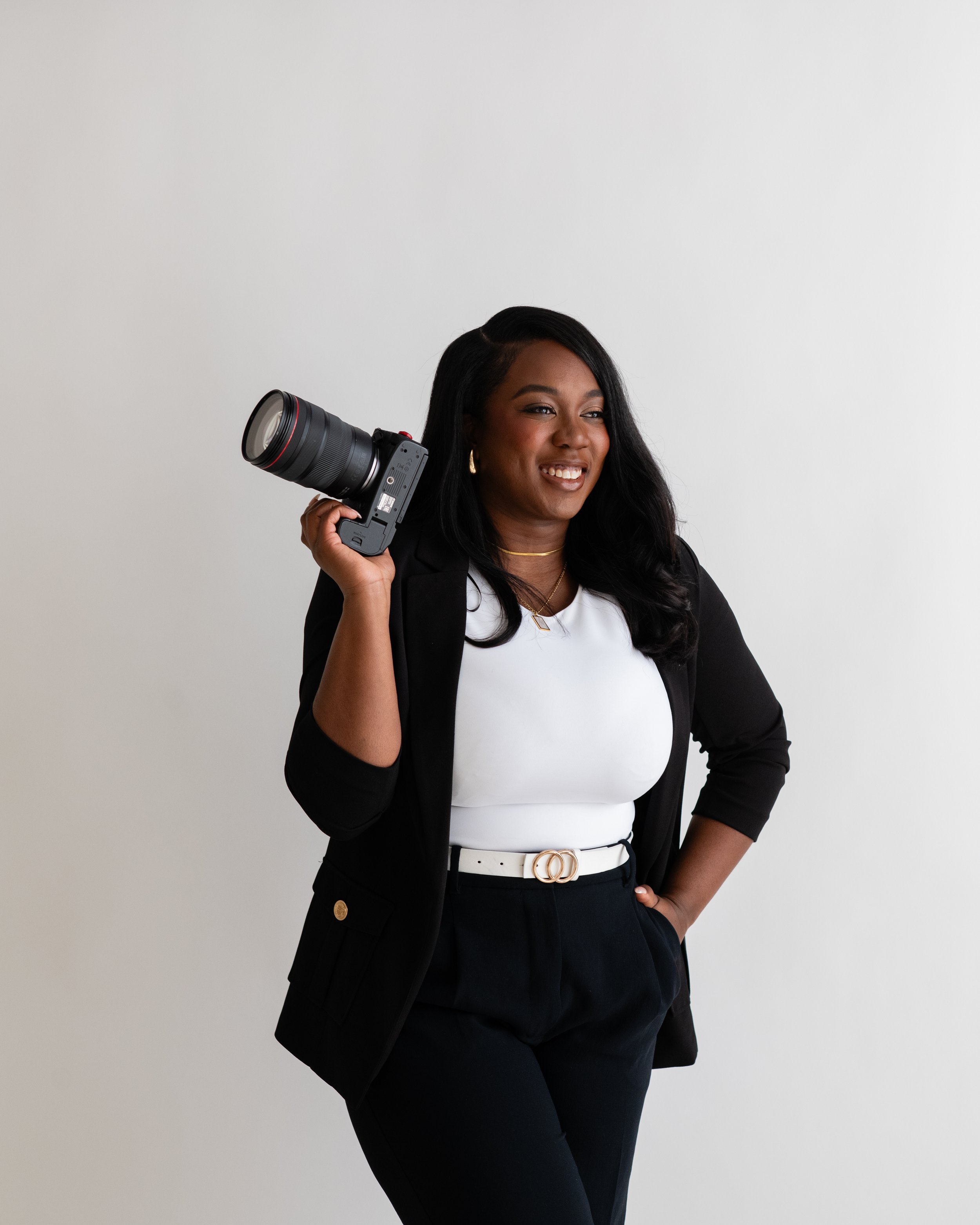 A woman in professional attire holding a camera over her shoulder, smiling in front of a plain white background.