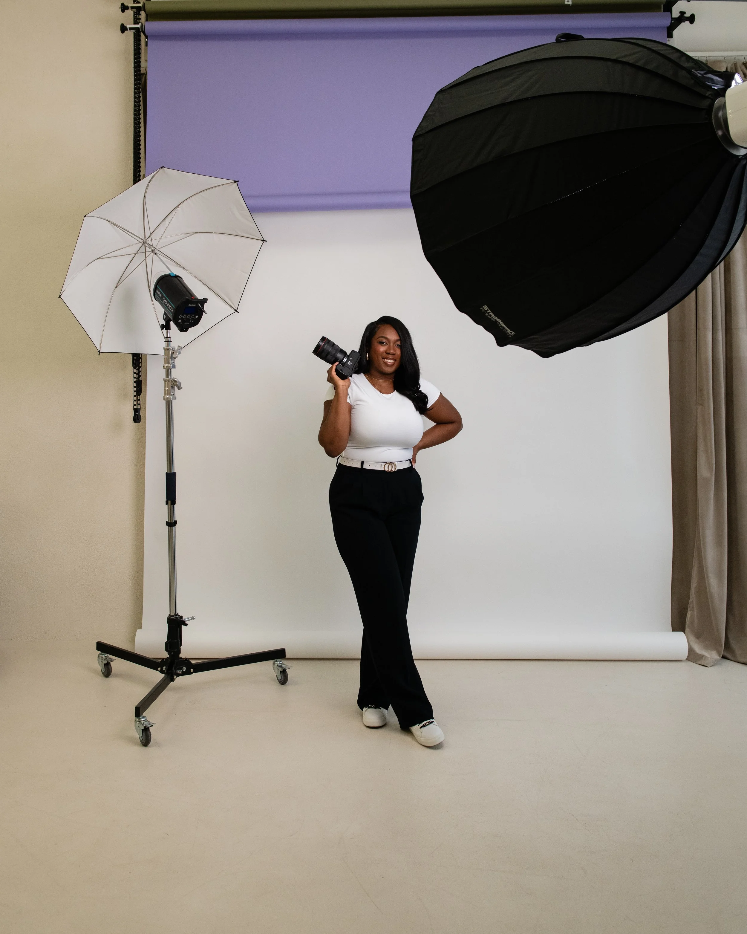 A woman standing in a photography studio, holding a camera, smiling, with studio lights and equipment around her, and a purple backdrop behind her.
