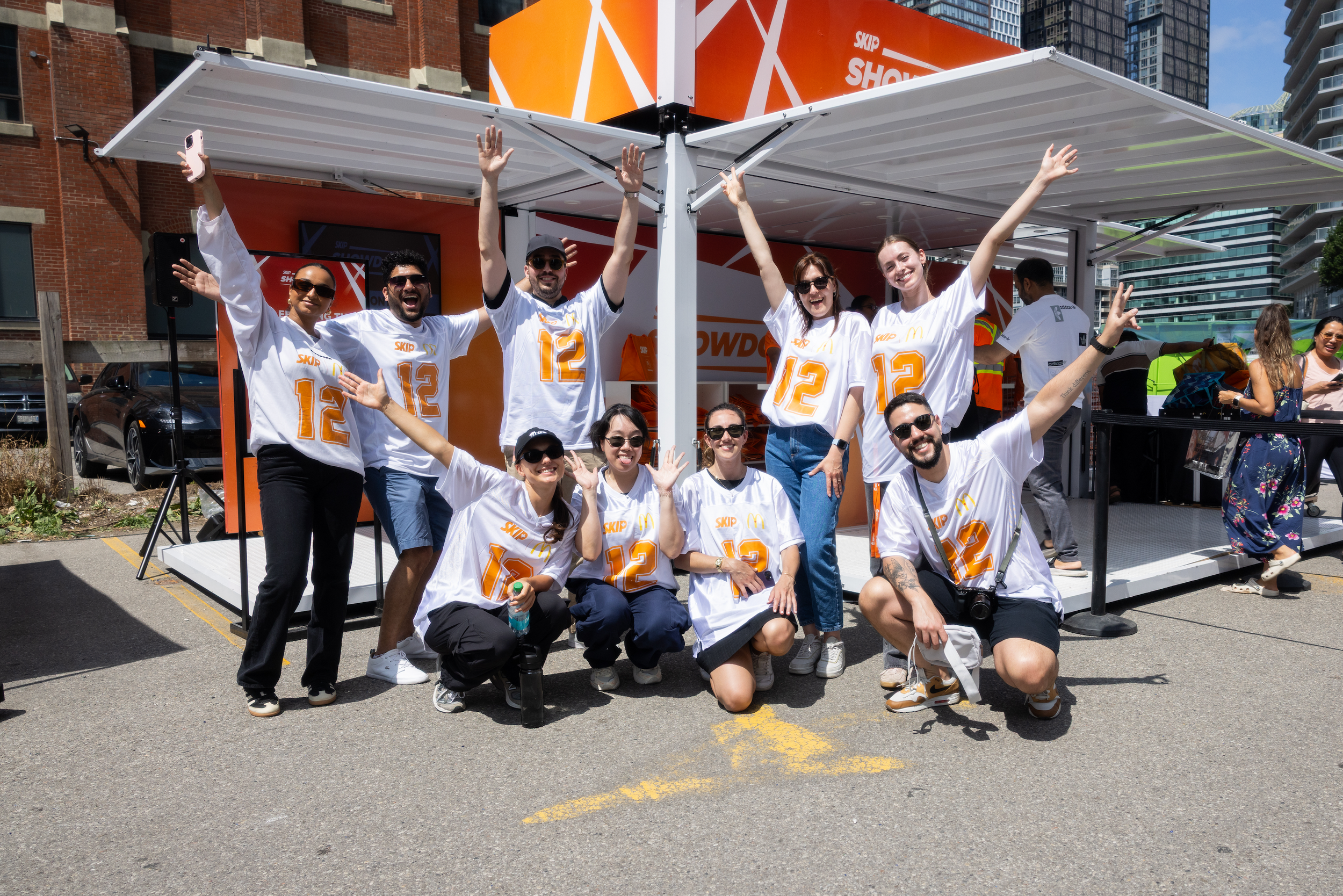 Group of nine people wearing matching white shirts with orange and yellow designs, posing cheerfully outside on a sunny day.