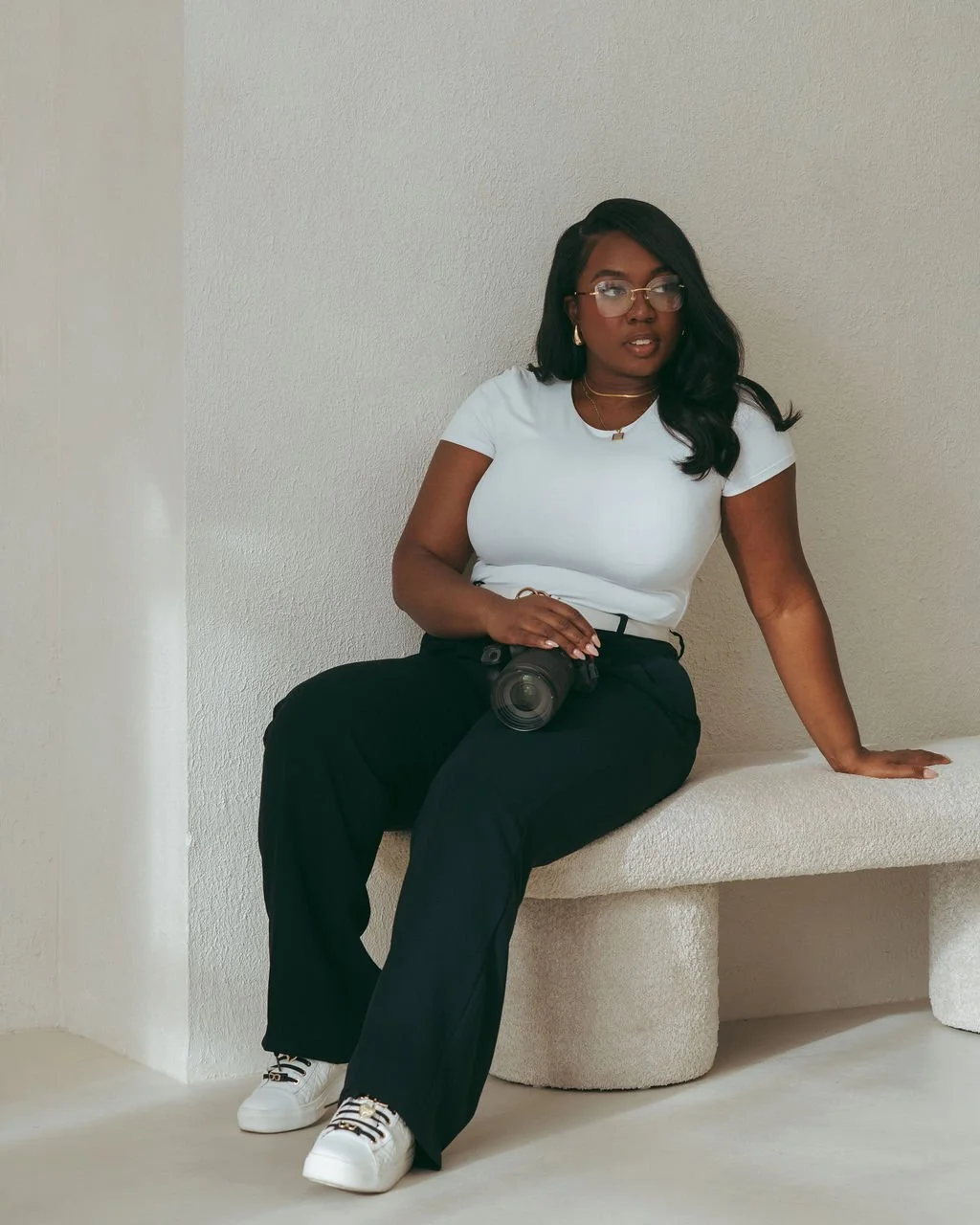 A woman with dark hair, glasses, and earrings sitting on a cream-colored bench against a plain white wall, holding a camera in her lap, wearing a white t-shirt, black pants, and white sneakers.