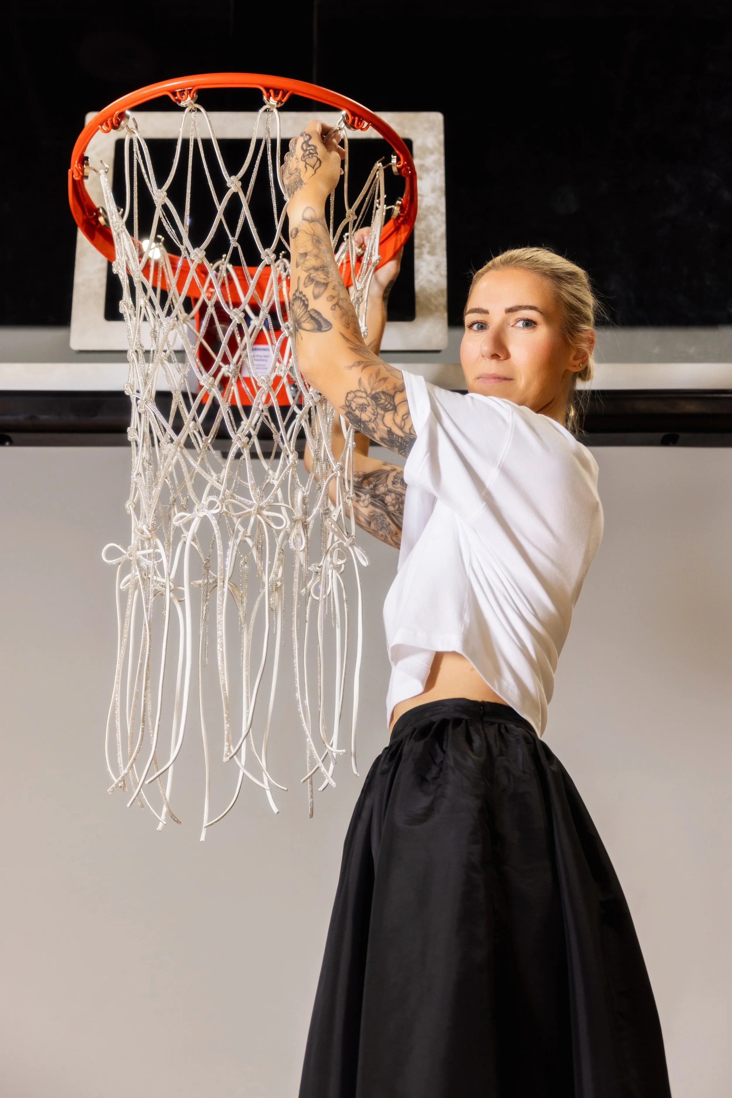 A woman with tattoos wearing a white t-shirt and black shorts is hanging a basketball net onto a hoop in a gym.